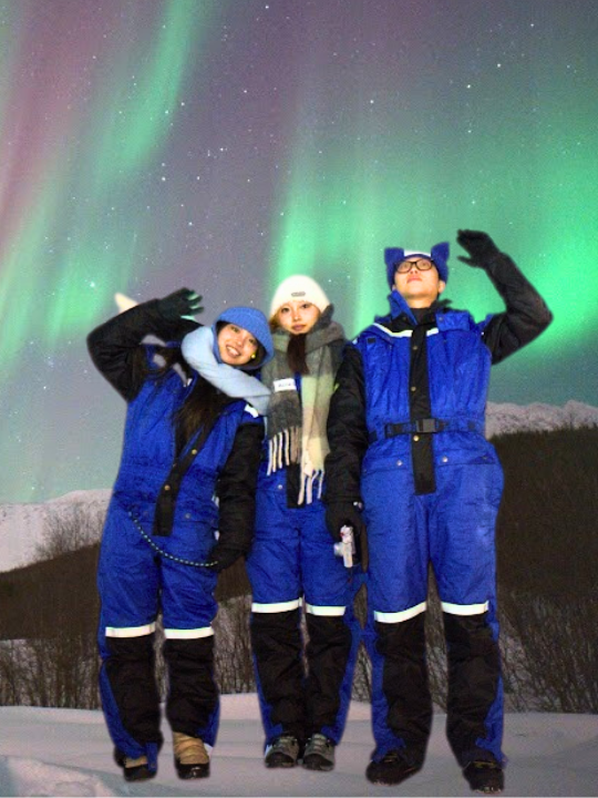 one man waving at he sky colored of aurora colors and two girls standing next to him dressed in thermal clothes