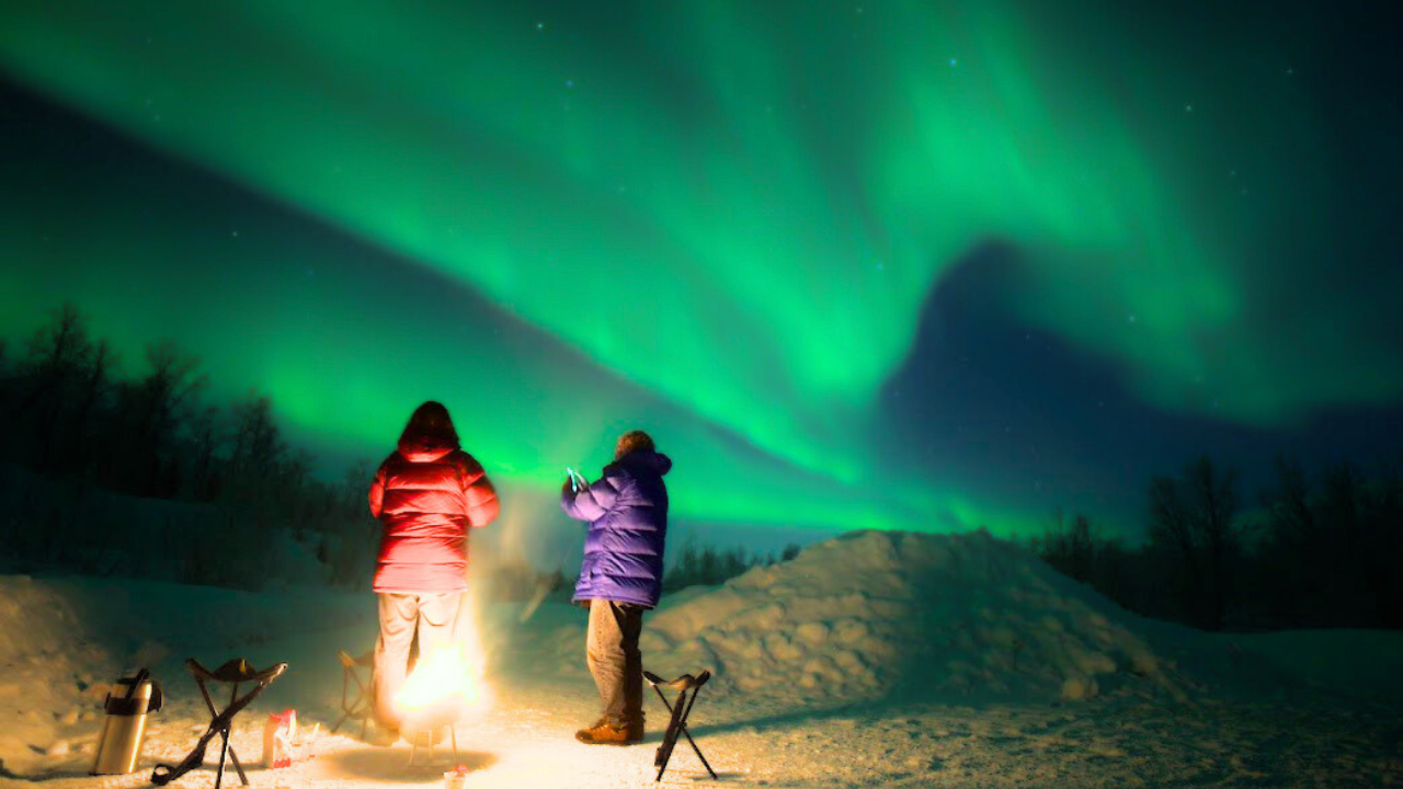 couples standing infront of camp fire under aurora sky