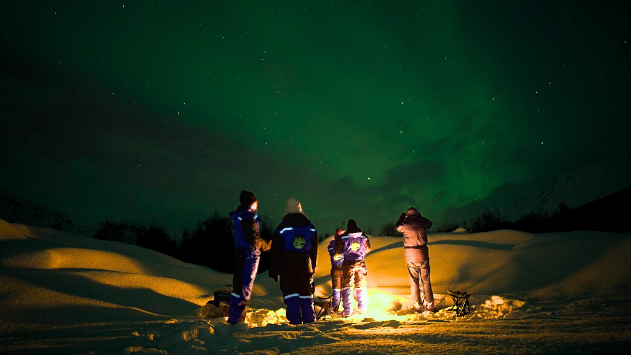 group of travelers standing infron of campfire under aurora sky ad snowy landscape
