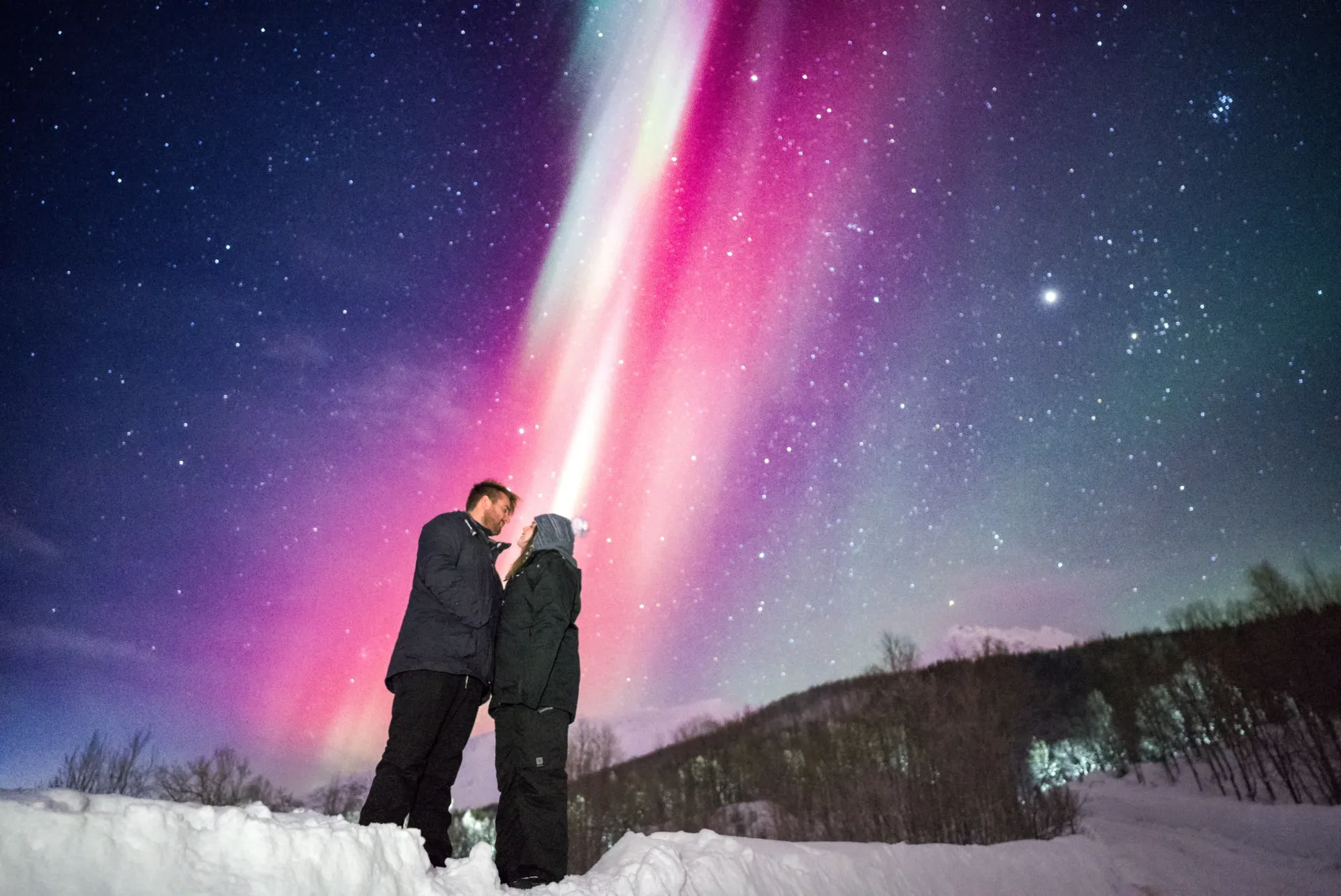 Couple standing under a vivid pink aurora display and starry night sky during an Alshemal Norway chase