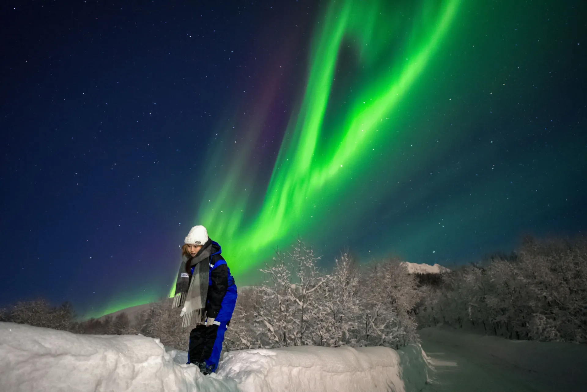 Traveler wearing a white beanie and Regatta thermal suit standing in snow beneath a green and purple Northern Lights curtain in Tromsø