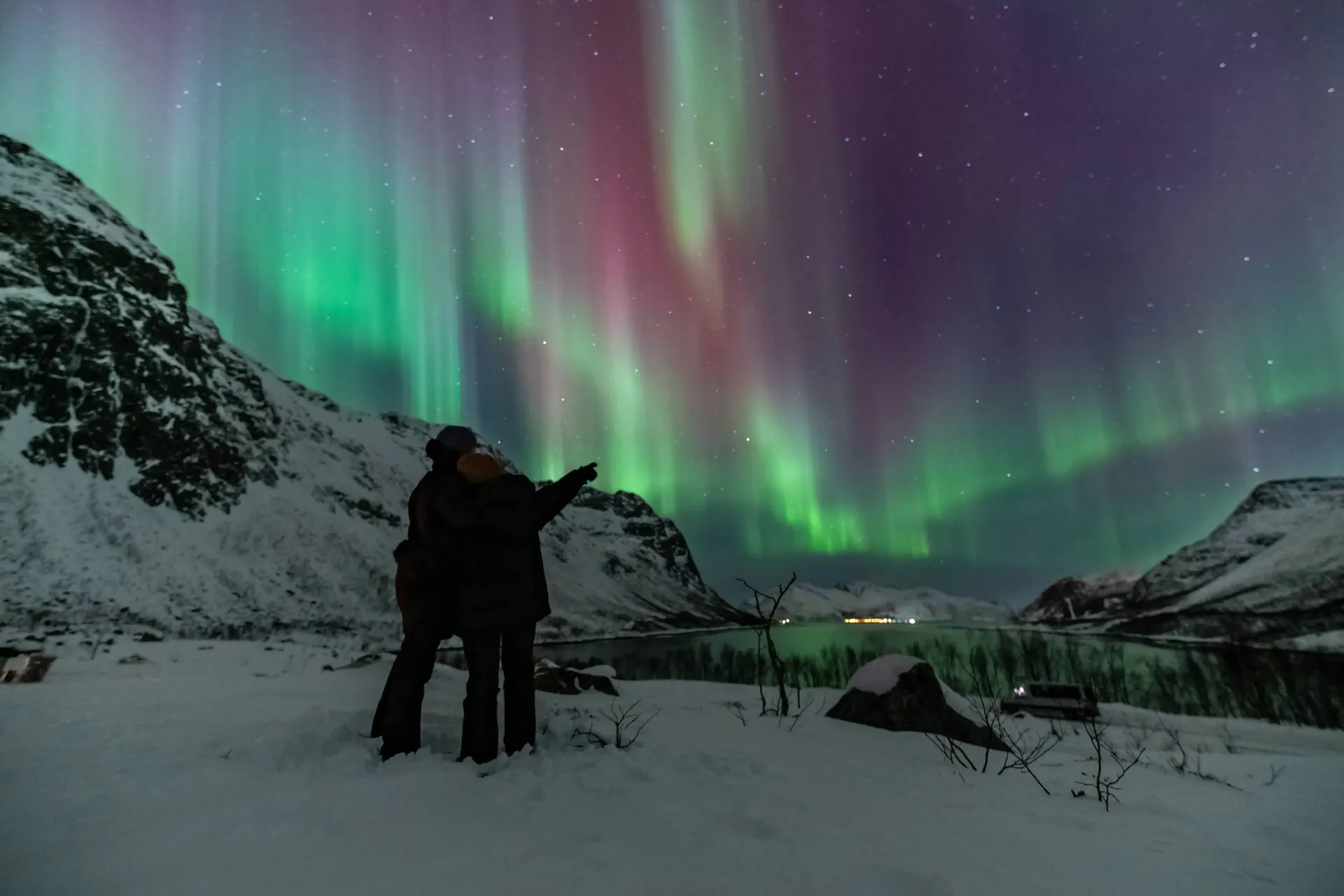 Small group of travelers in thermal suits watching bright green Northern Lights over snow-covered Arctic landscape in Tromsø