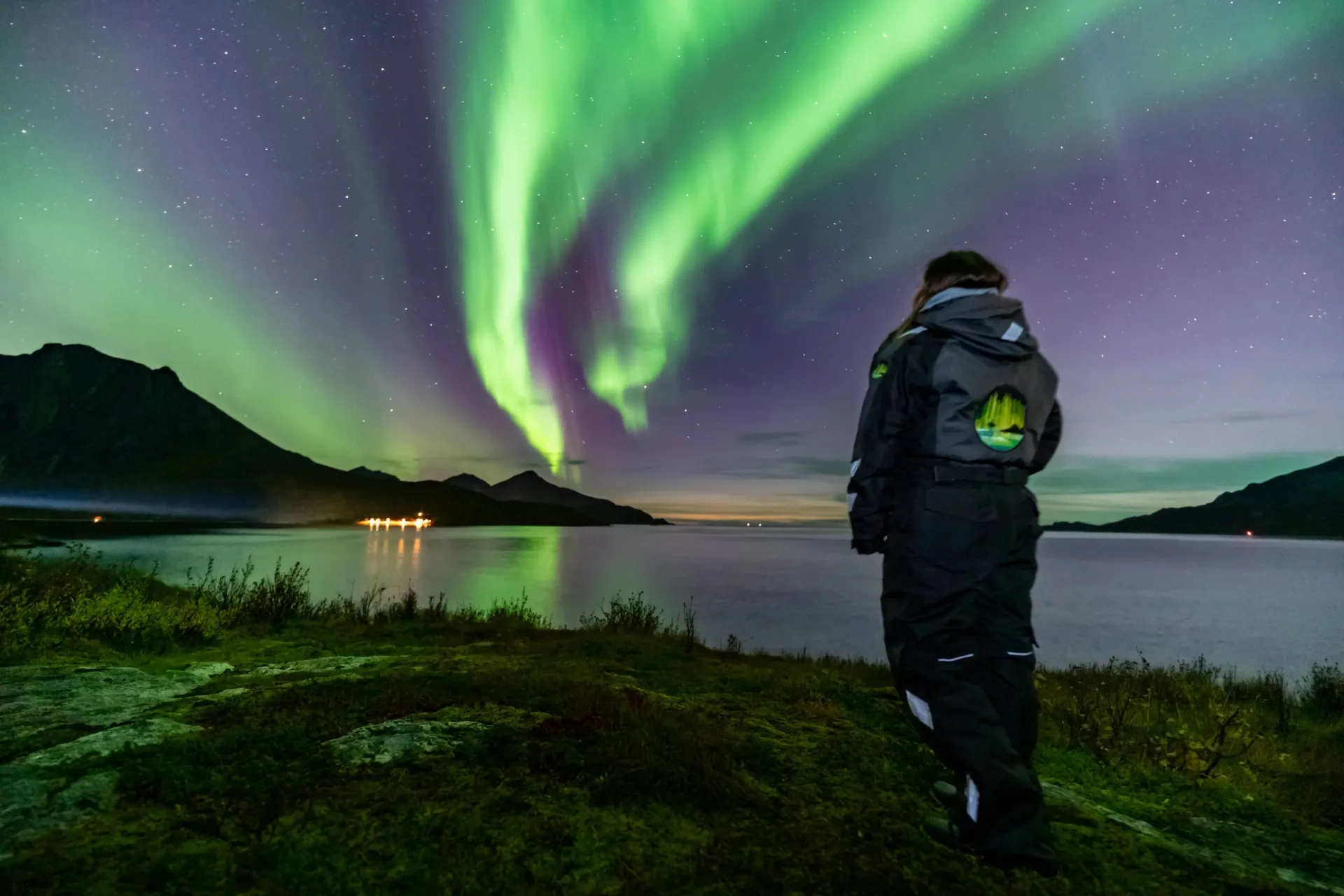Alshemal guide in thermal suit standing on coastal shoreline looking at green and purple Northern Lights over Tromsø fjord