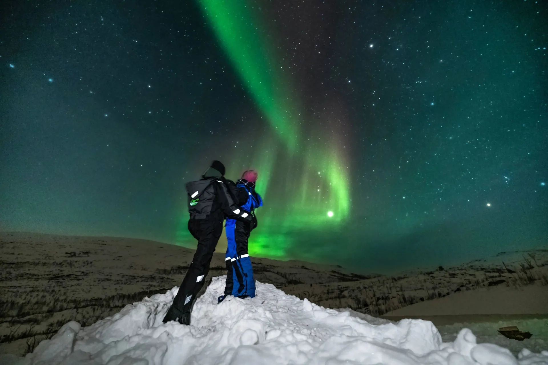 Two guests in thermal suits standing on a snowy ridge under a brilliant green Northern Lights beam in Tromsø