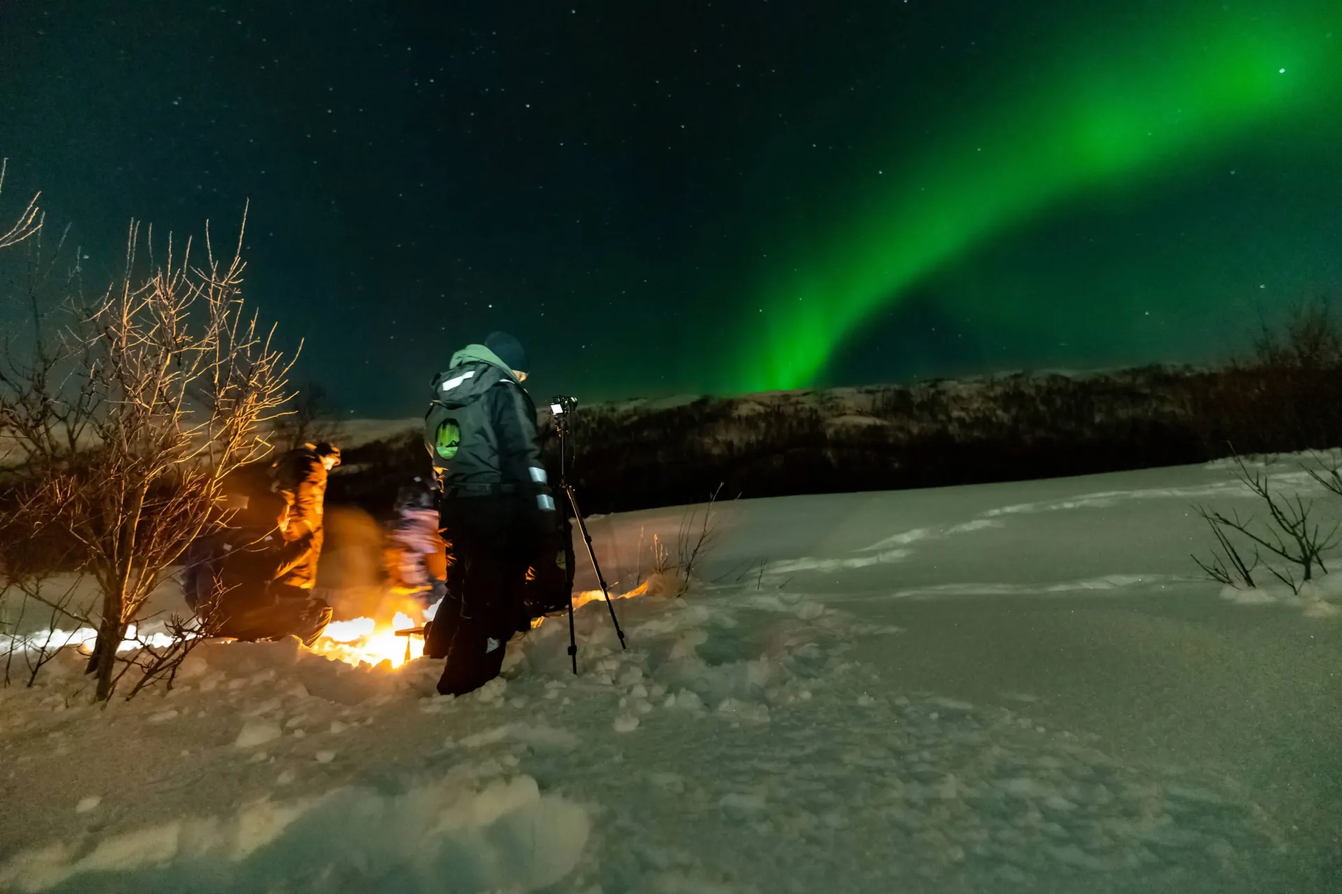 Guests in thermal suits around a campfire in deep snow with camera tripod under green Northern Lights