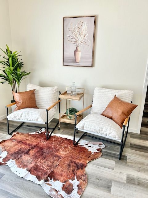 Two white chairs, leather pillows, wooden side table, cowhide rug, and framed art in a neutral-toned room.