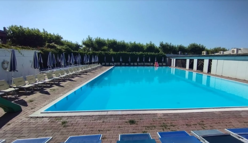 Una lunga piscina rettangolare con acqua di un azzurro brillante, circondata da file di ombrelloni e lettini su un patio.