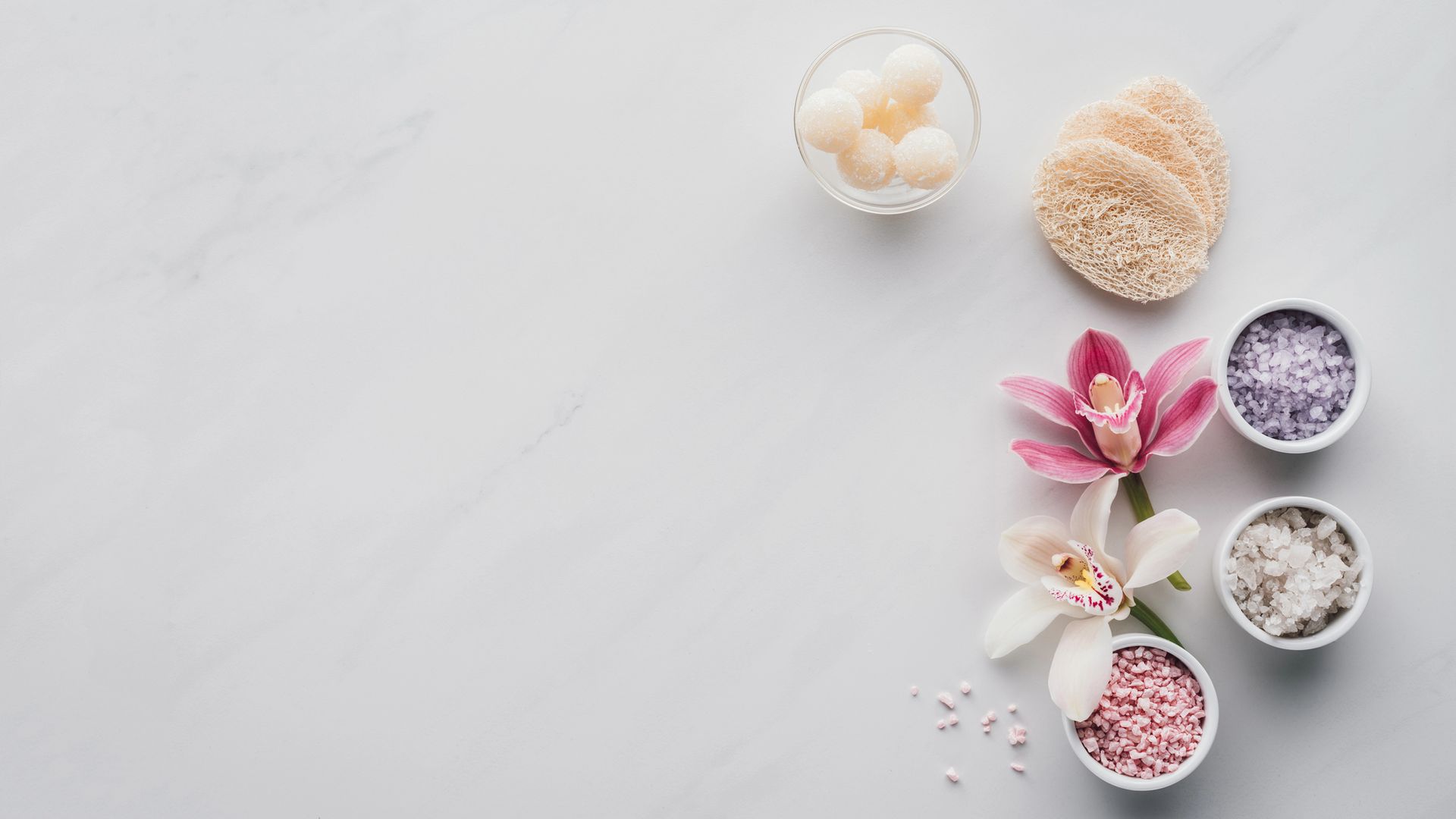 Articoli per la spa su uno sfondo grigio chiaro: spugne vegetali, sali da bagno in piccole ciotole e fiori di orchidea rosa e bianchi.