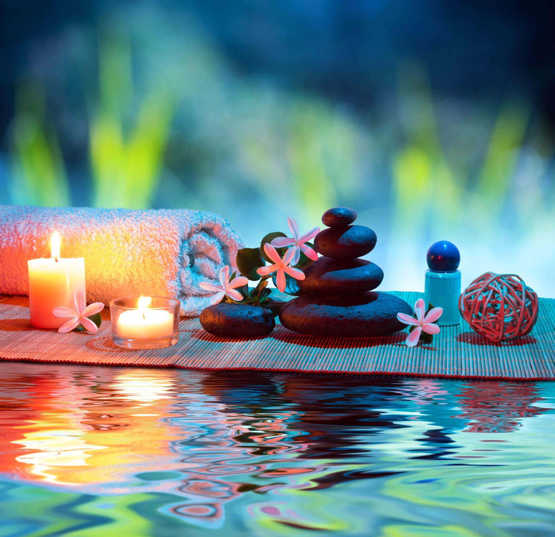 Ambientazione da spa con candele accese, pietre impilate, asciugamano e fiori che si riflettono sull'acqua su uno sfondo verde sfocato.