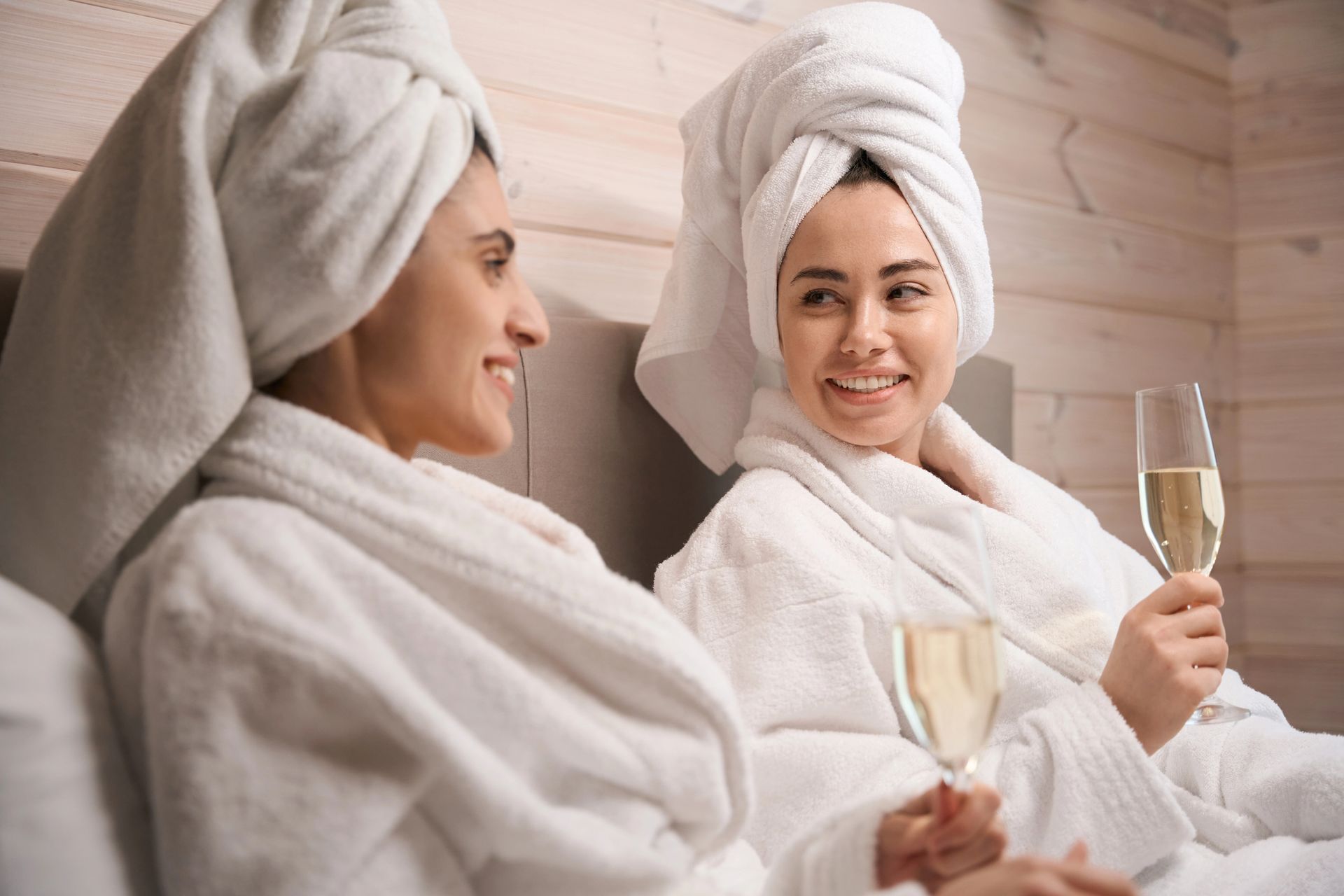 In una stanza con pareti in legno, due persone in accappatoio e con asciugamani per capelli siedono insieme, sorridenti e con in mano calici di champagne.