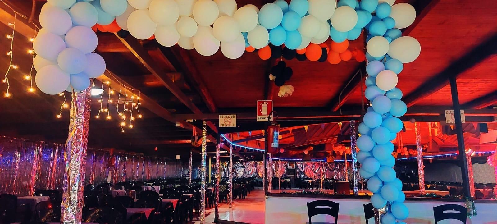 Un ampio spazio per eventi al coperto con un arco di palloncini nei toni del blu e del bianco, luci decorative e un'illuminazione d'atmosfera sui toni del rosso.