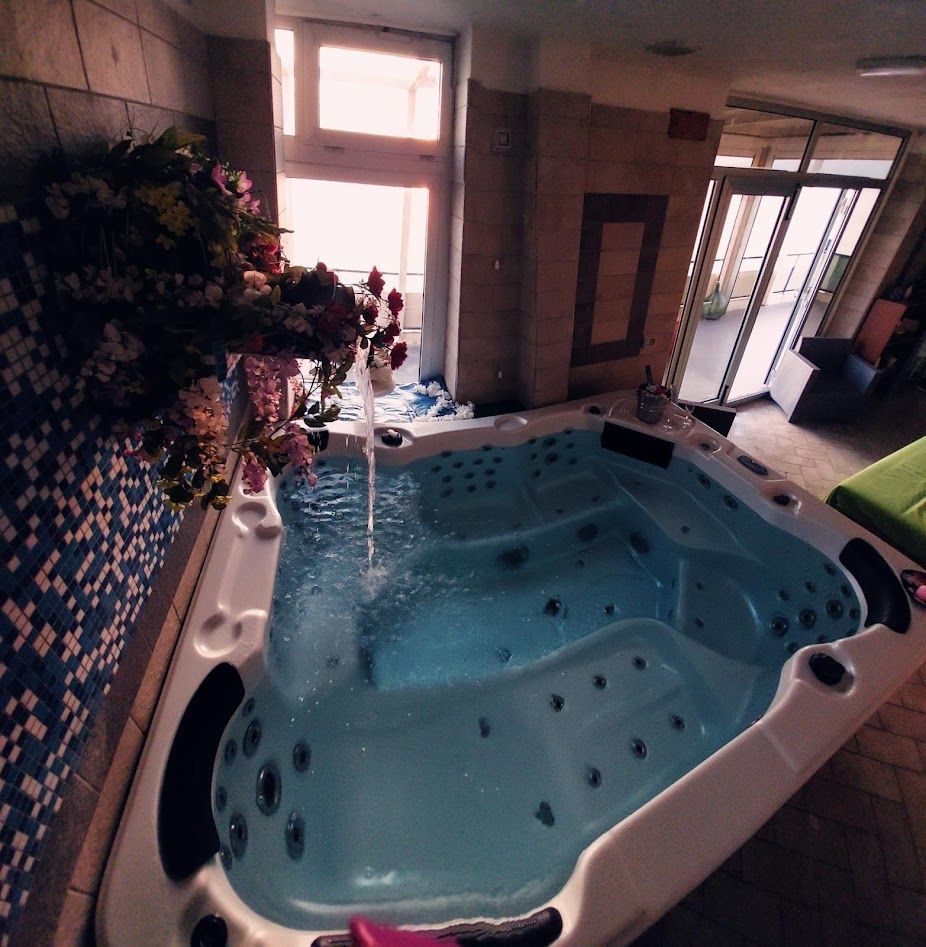 Una grande vasca idromassaggio azzurra si riempie d'acqua sotto una fontana decorativa a forma di fiori, all'interno di una stanza con pareti e finestre piastrellate.