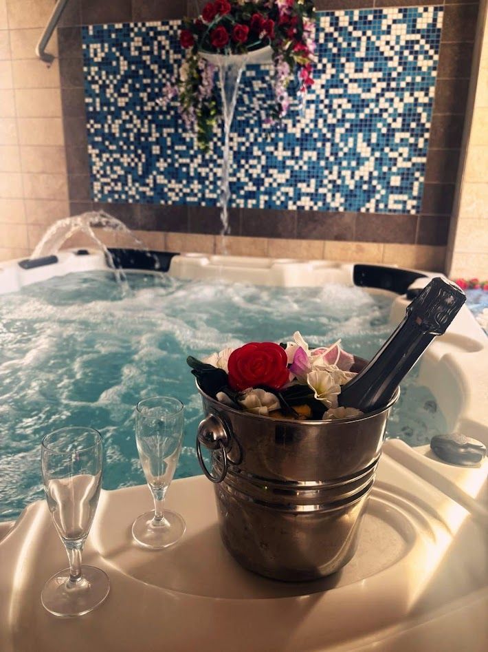 Un secchiello di metallo per champagne decorato con rose e una bottiglia si trova accanto a una vasca idromassaggio gorgogliante, con due bicchieri appoggiati su un ripiano piastrellato.