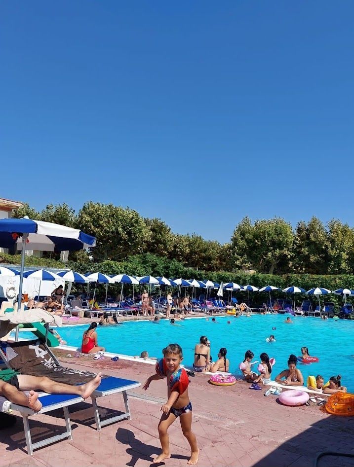 Una soleggiata piscina all'aperto con lettini, ombrelloni blu e bianchi e molte persone che nuotano o si rilassano.