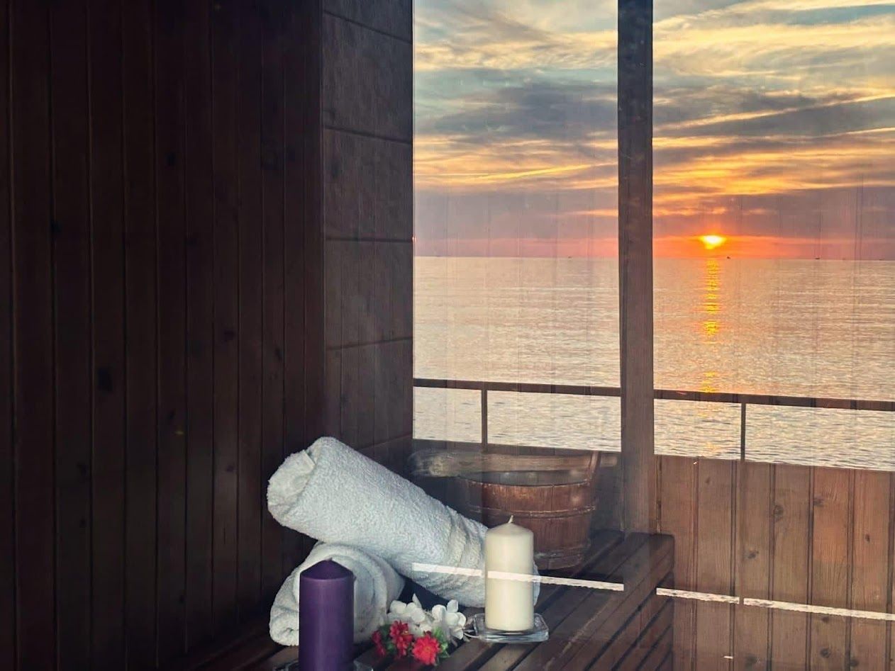 Due asciugamani arrotolati, una candela e dei piccoli fiori su una superficie di legno con vista su un tramonto sull'oceano.