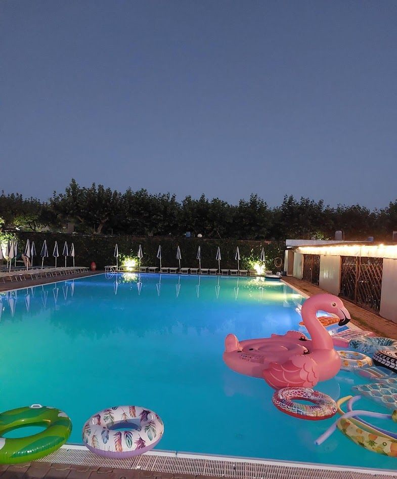 Una piscina di un blu intenso al crepuscolo, con diversi giochi gonfiabili colorati, tra cui un grande fenicottero rosa.