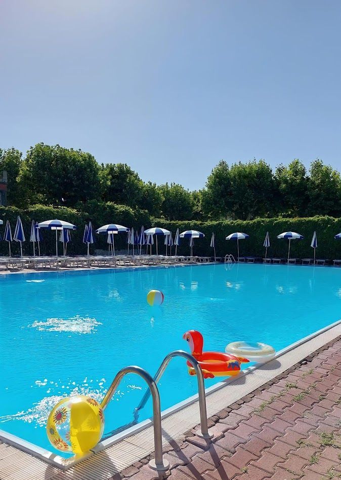 Una piscina di un blu brillante con colorati giochi gonfiabili, ombrelloni a bordo piscina e alberi rigogliosi sotto un cielo limpido.