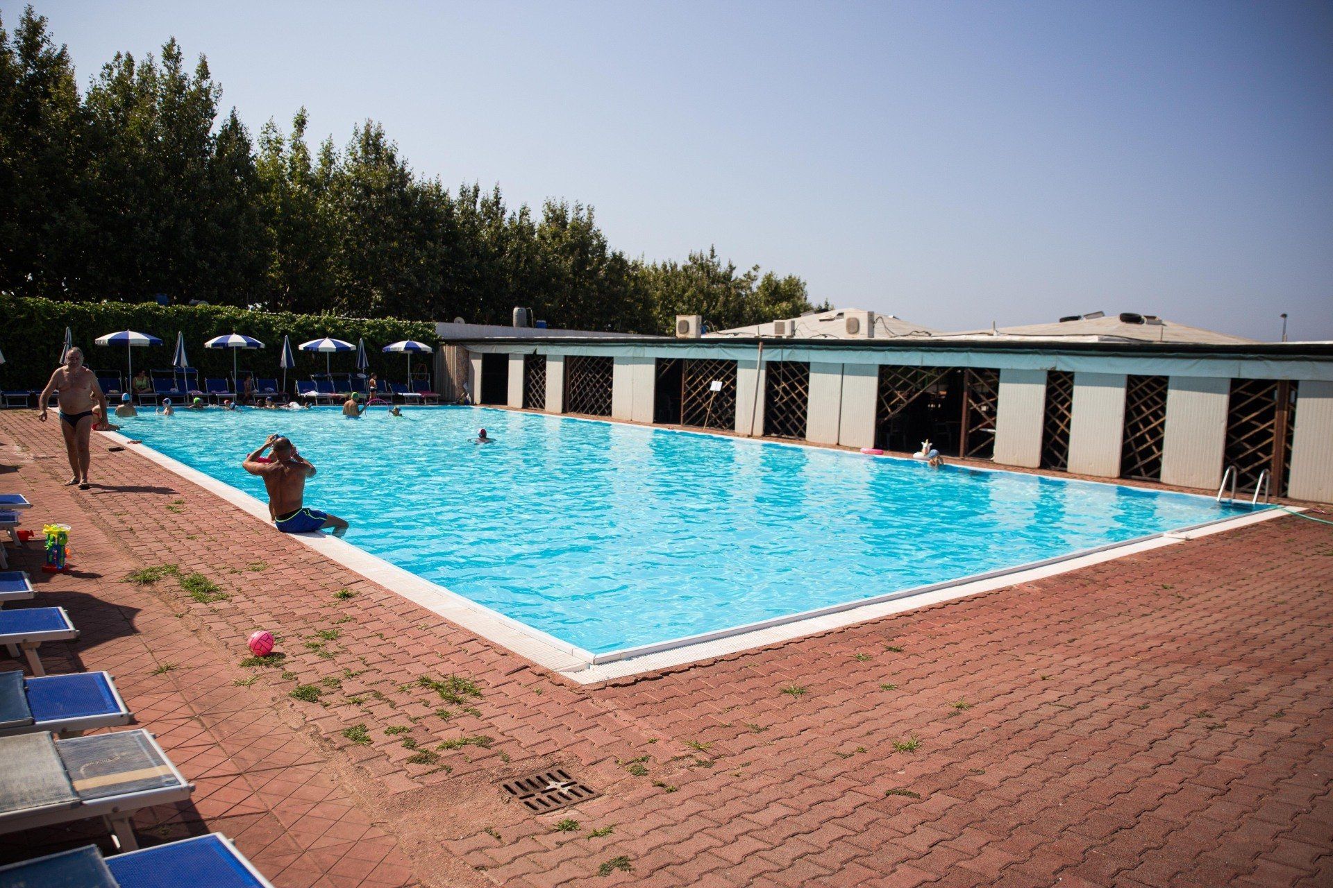 Persone che nuotano in una piscina rettangolare all'aperto con un pavimento in mattoni rossi, circondata da alberi e da un edificio con vetrate aperte.