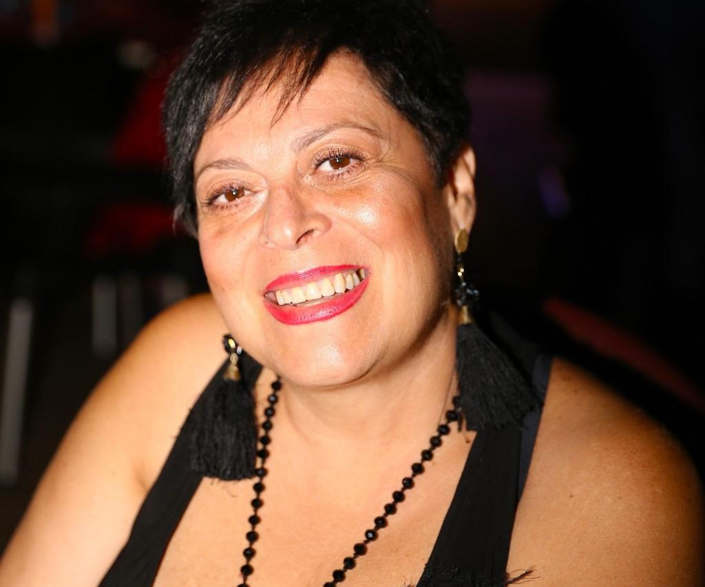 Una persona sorridente con capelli corti e scuri, che indossa una maglietta nera, orecchini con nappe e una collana di perline.