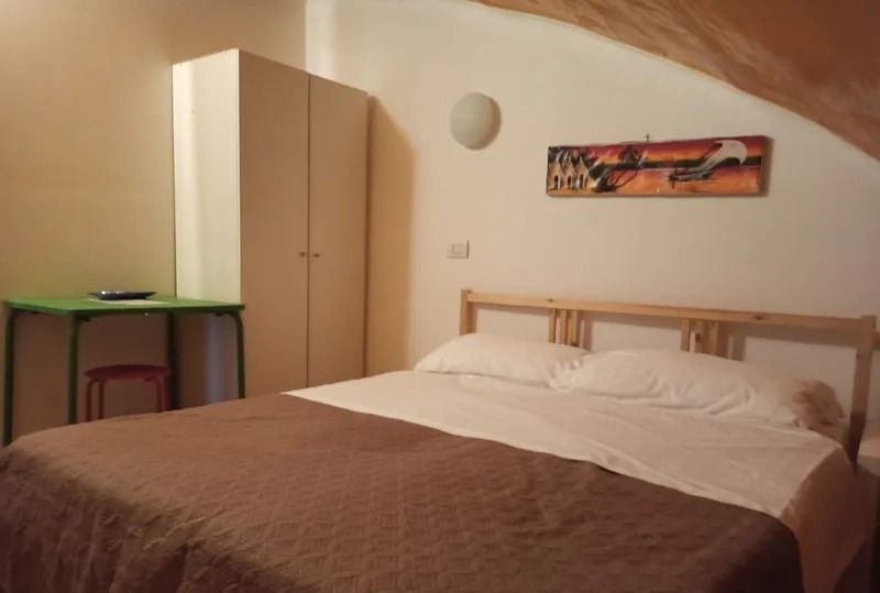 Una camera da letto con un letto matrimoniale, una trapunta marrone, cuscini bianchi, una testiera in legno, un armadio bianco e una scrivania verde con sgabello.
