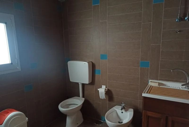 Un bagno con pareti rivestite di piastrelle marroni, un WC, un bidet e un mobiletto in legno con lavabo bianco e rubinetto in metallo.