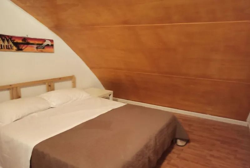 Una camera da letto con soffitto in legno inclinato, un letto con cuscini bianchi e una coperta marrone, e una decorazione a parete sulla sinistra.