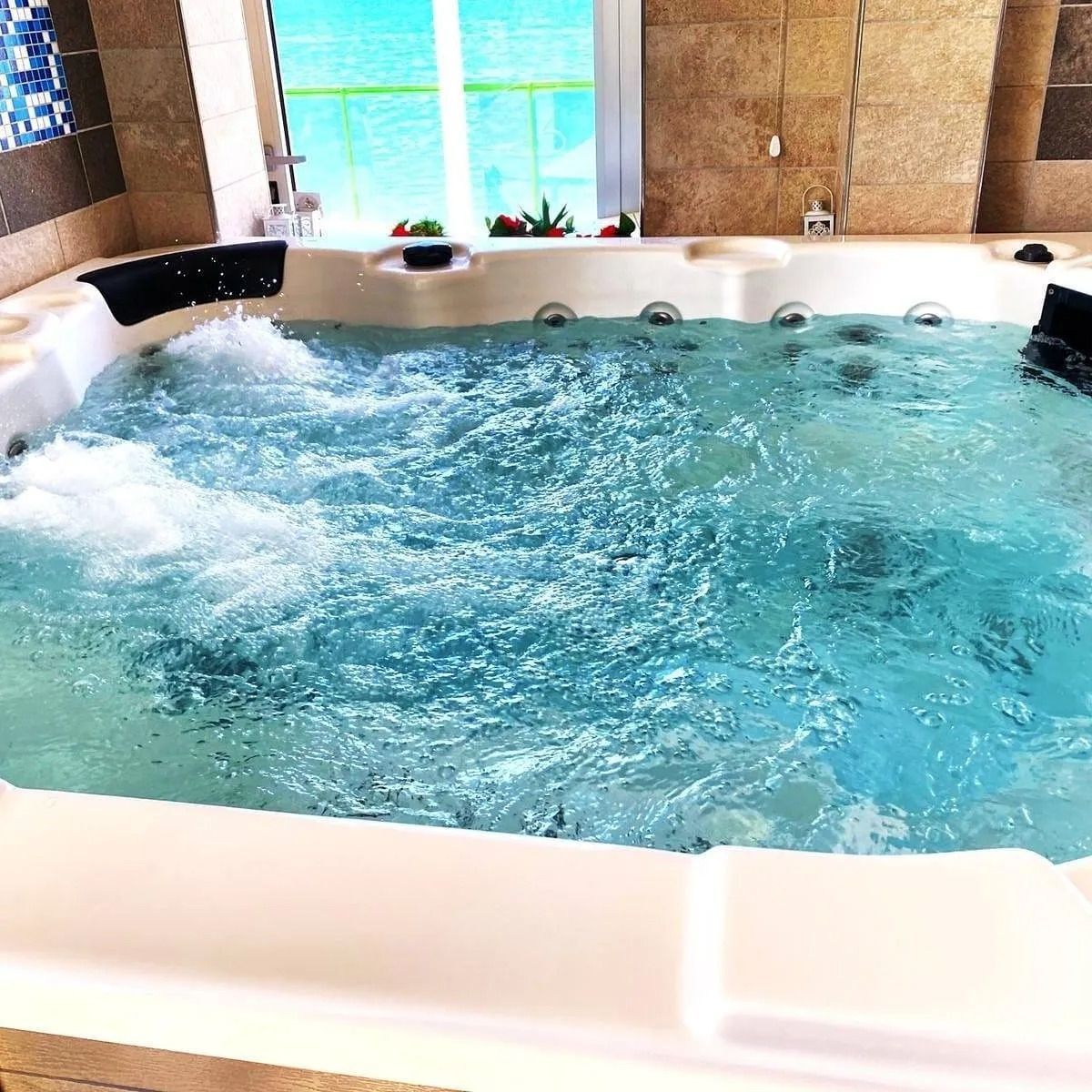 Una vasca idromassaggio bianca e quadrata, riempita di acqua blu gorgogliante, addossata a una parete rivestita di piastrelle di pietra vicino a una finestra.