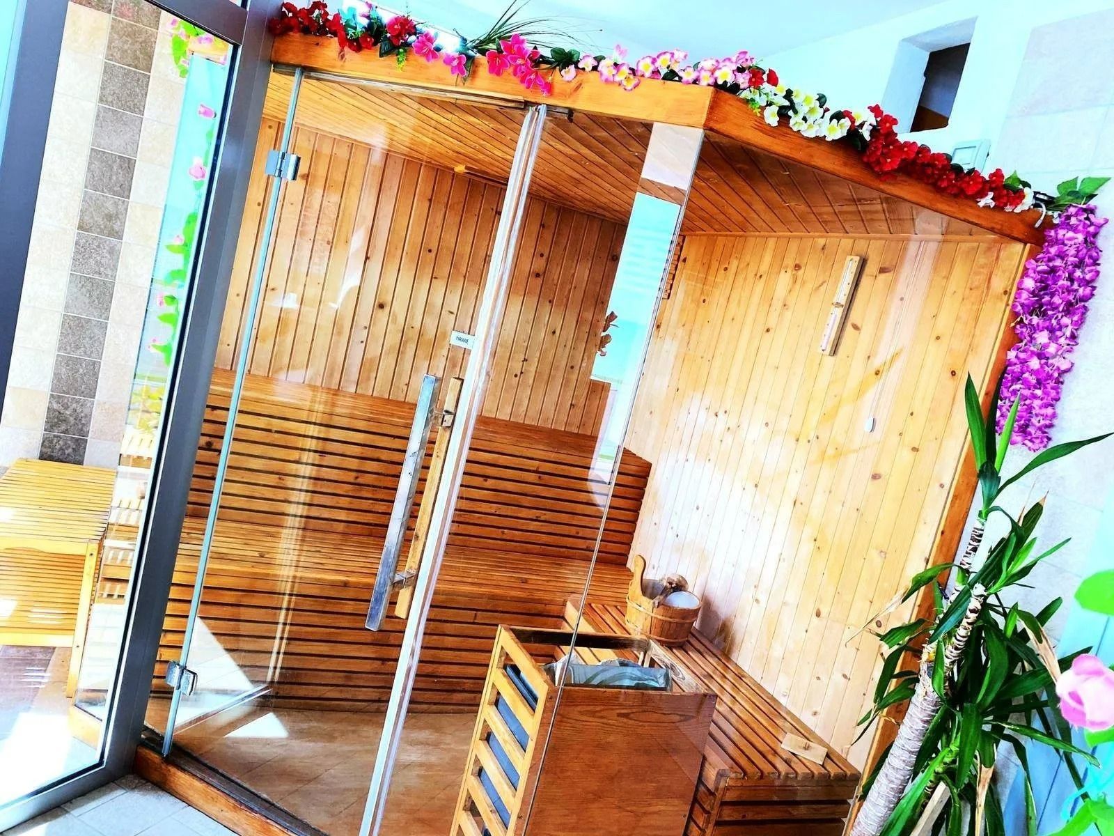 Una cabina sauna in legno con porte in vetro, decorata con ghirlande floreali, situata in un luminoso ambiente interno.