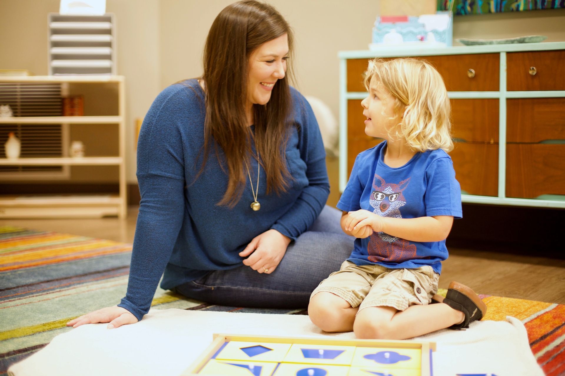 Adult and child seated on the floor, smiling and reading a book in a classroom.