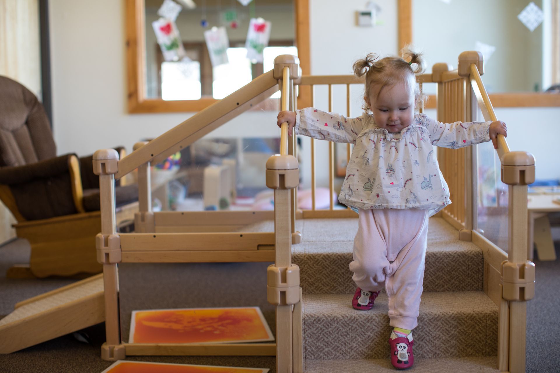 Child walking on a wooden play structure indoors, holding the railings, with a cozy living room in the background