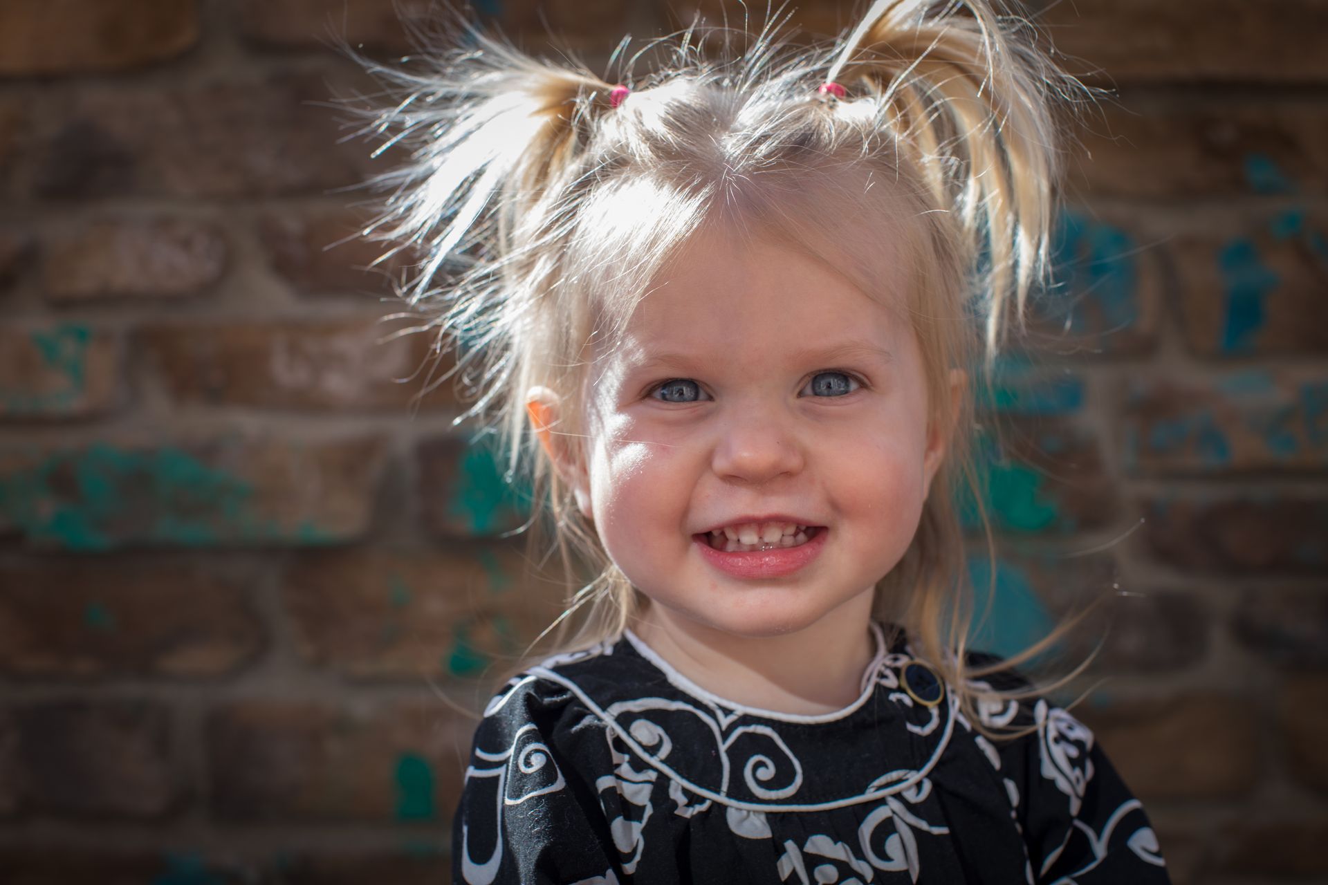 Smiling child with blonde hair in pigtails, wearing a black patterned shirt, standing before a brick wall.