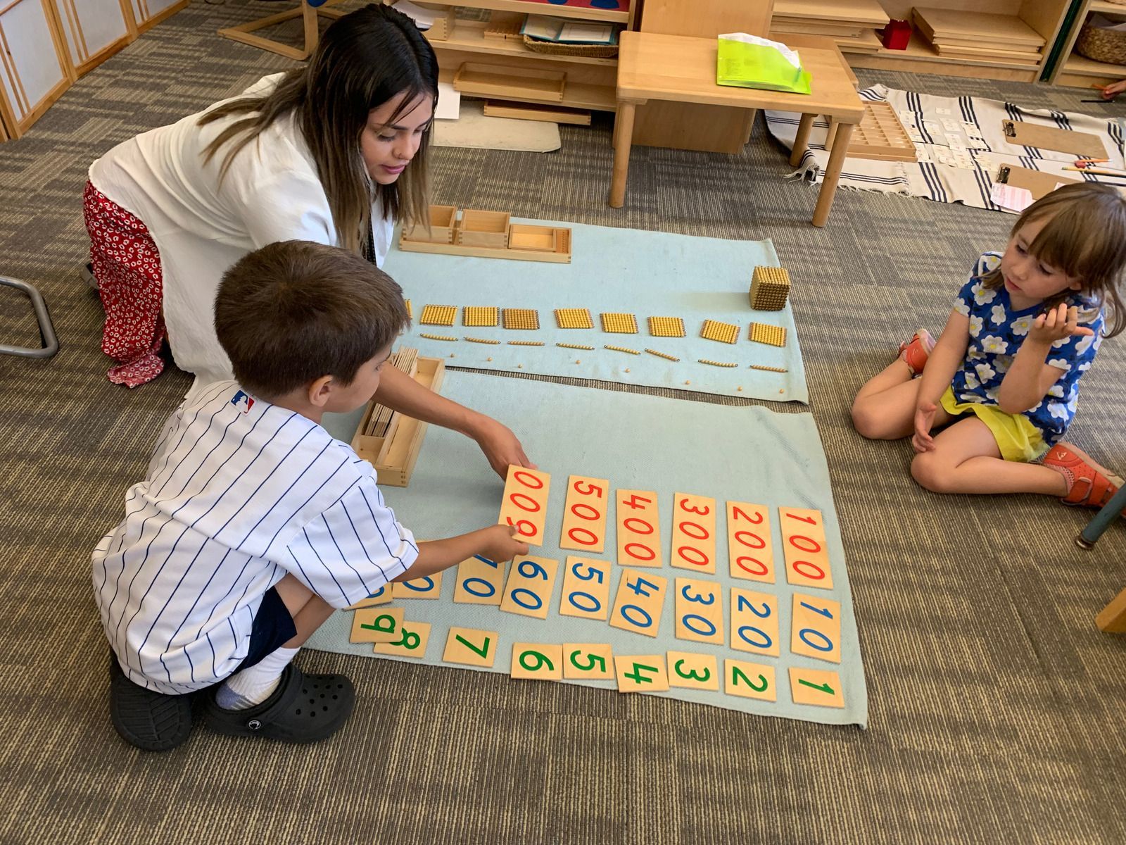 A montessori guide is showing two children a montessori math material with numbers.