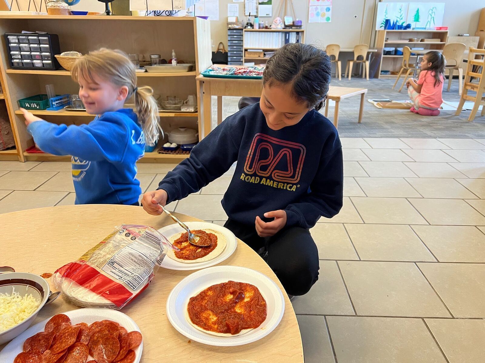 Two young girls are sitting at a table making pizza.