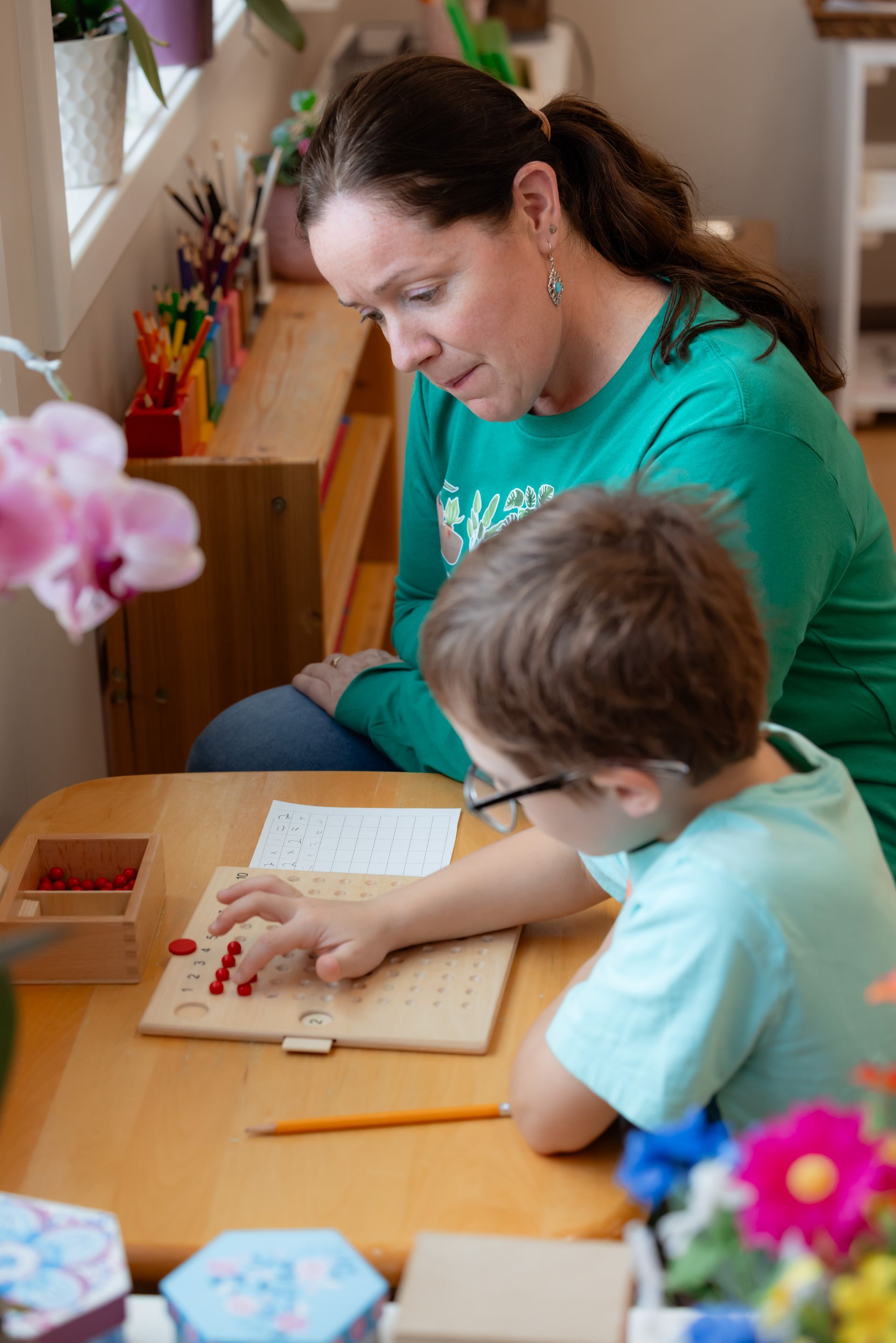 Adult and child working together at a table with colored pencils and paper, near a window