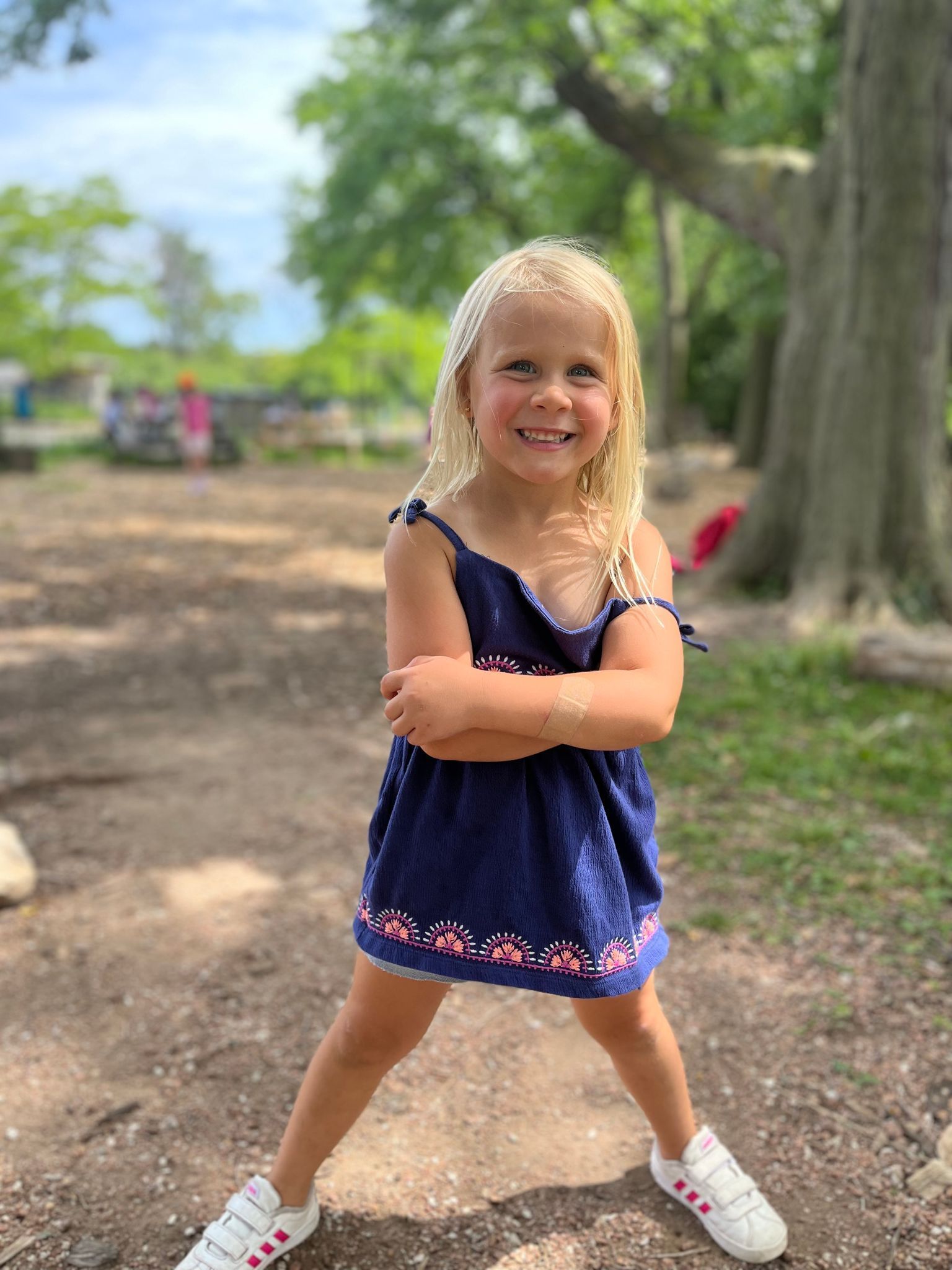 A little girl in a blue dress and white sneakers is standing on the ground with her arms crossed.