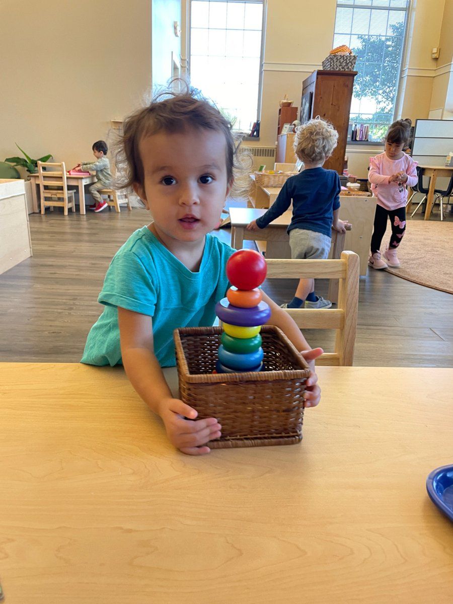 A little girl is sitting at a table holding a basket of montessori materials.
