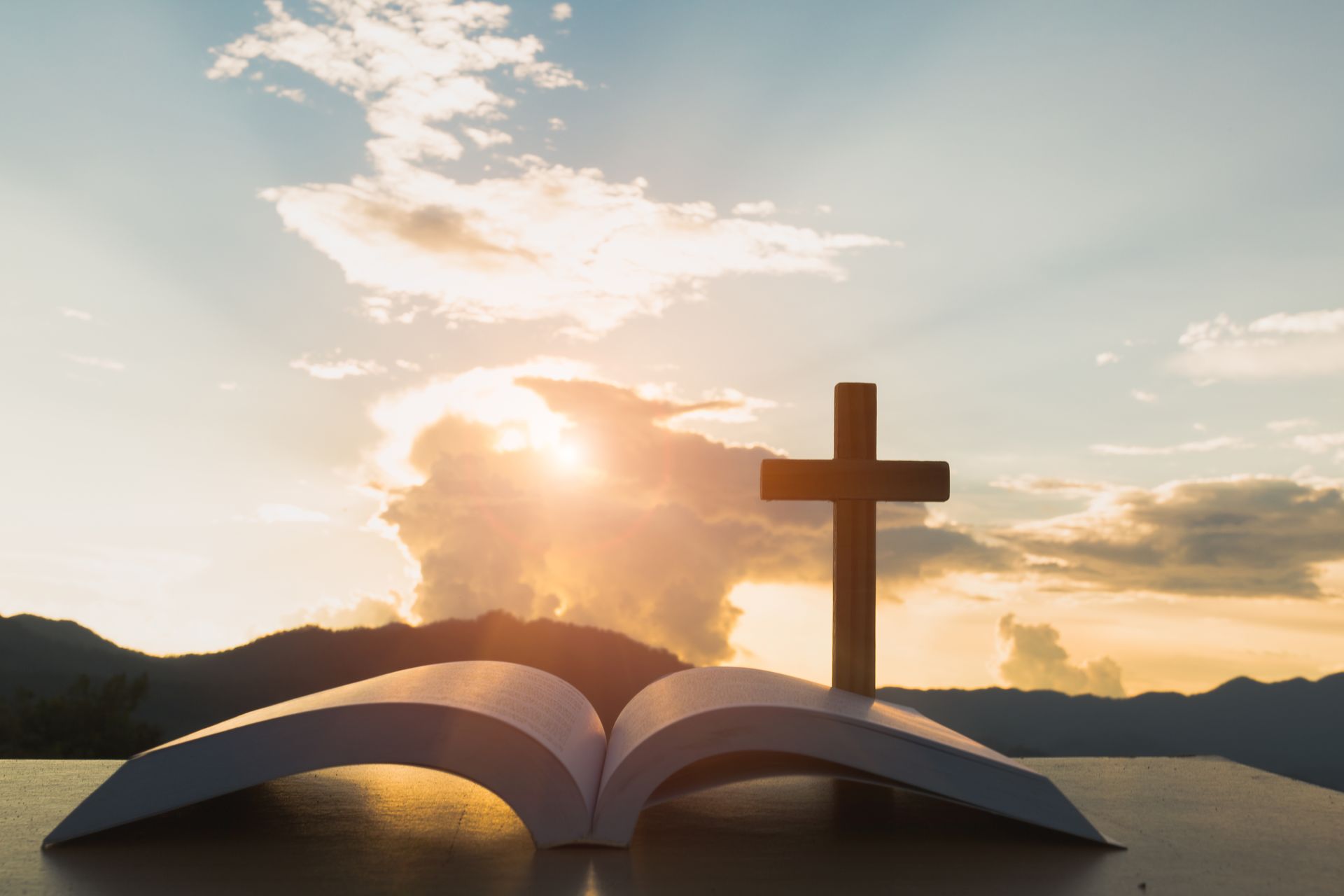 Open Bible and cross silhouetted against a sunset with mountains.