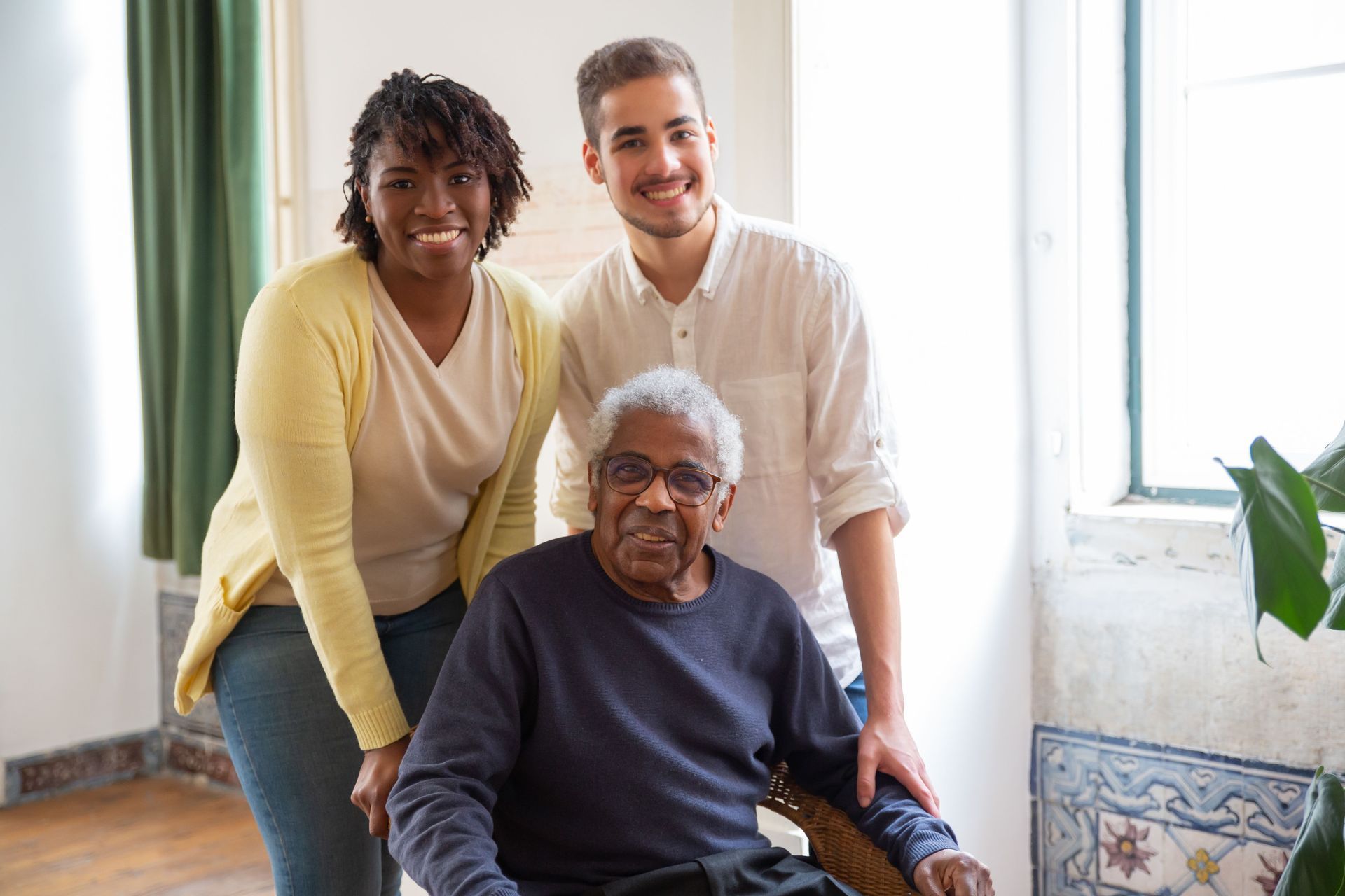 Tres personas sonriendo en una luminosa sala de estar; una de ellas está sentada y las otras dos están de pie a su lado.