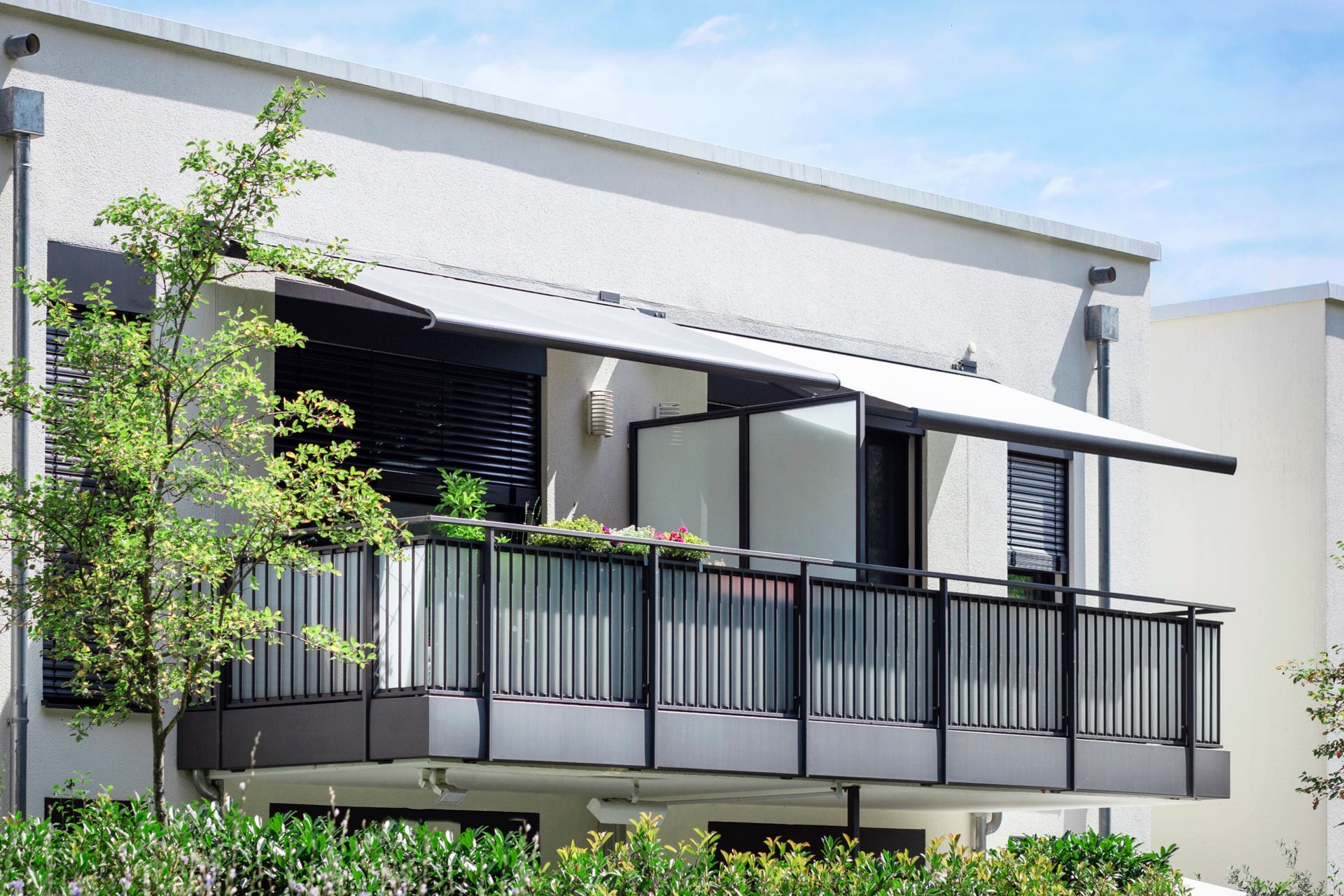 Modern apartment balcony with black railing, awning, and potted plants under a bright sky