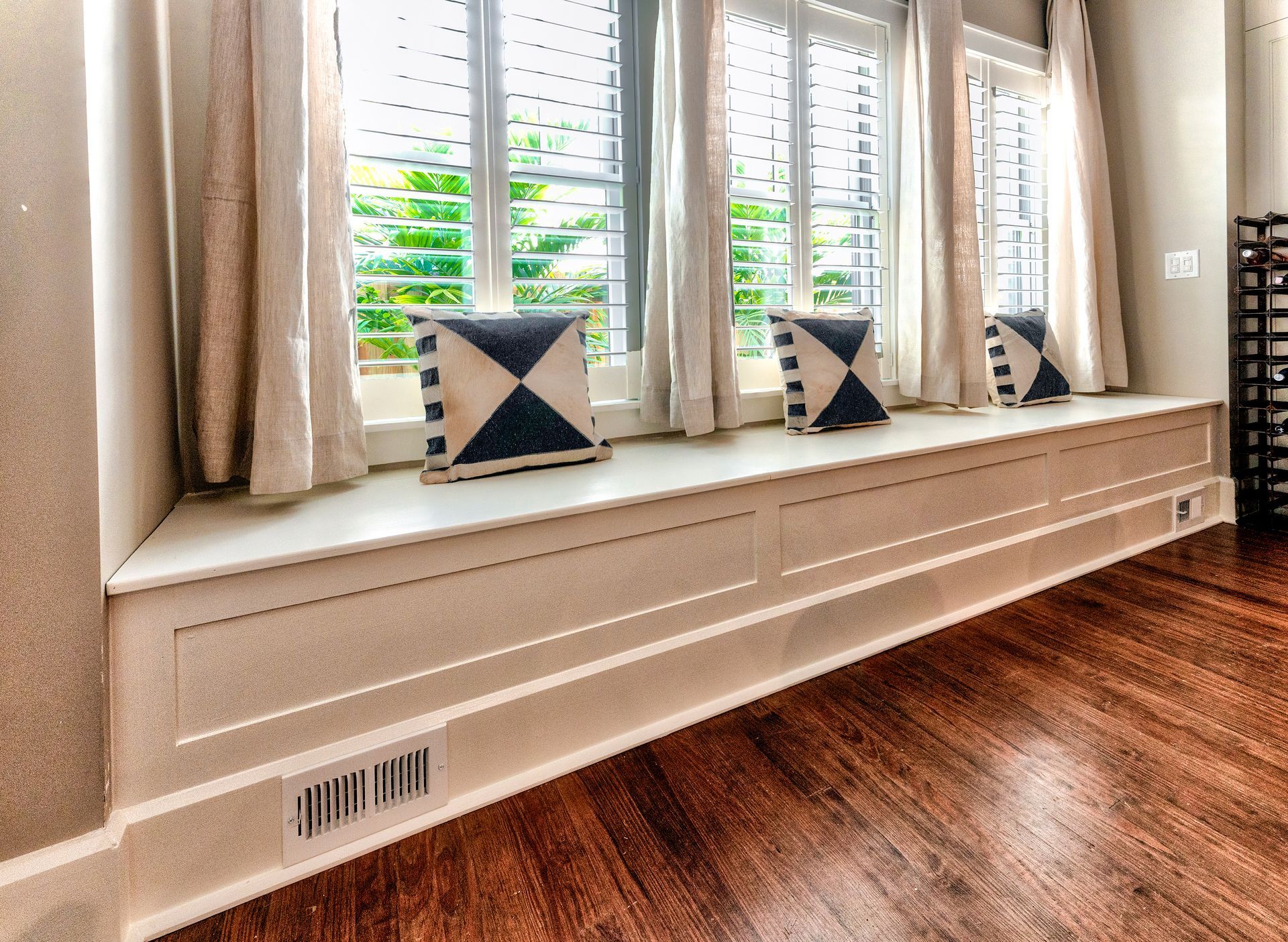Window seat with white cushion, patterned pillows, curtains, and hardwood floor in a bright room