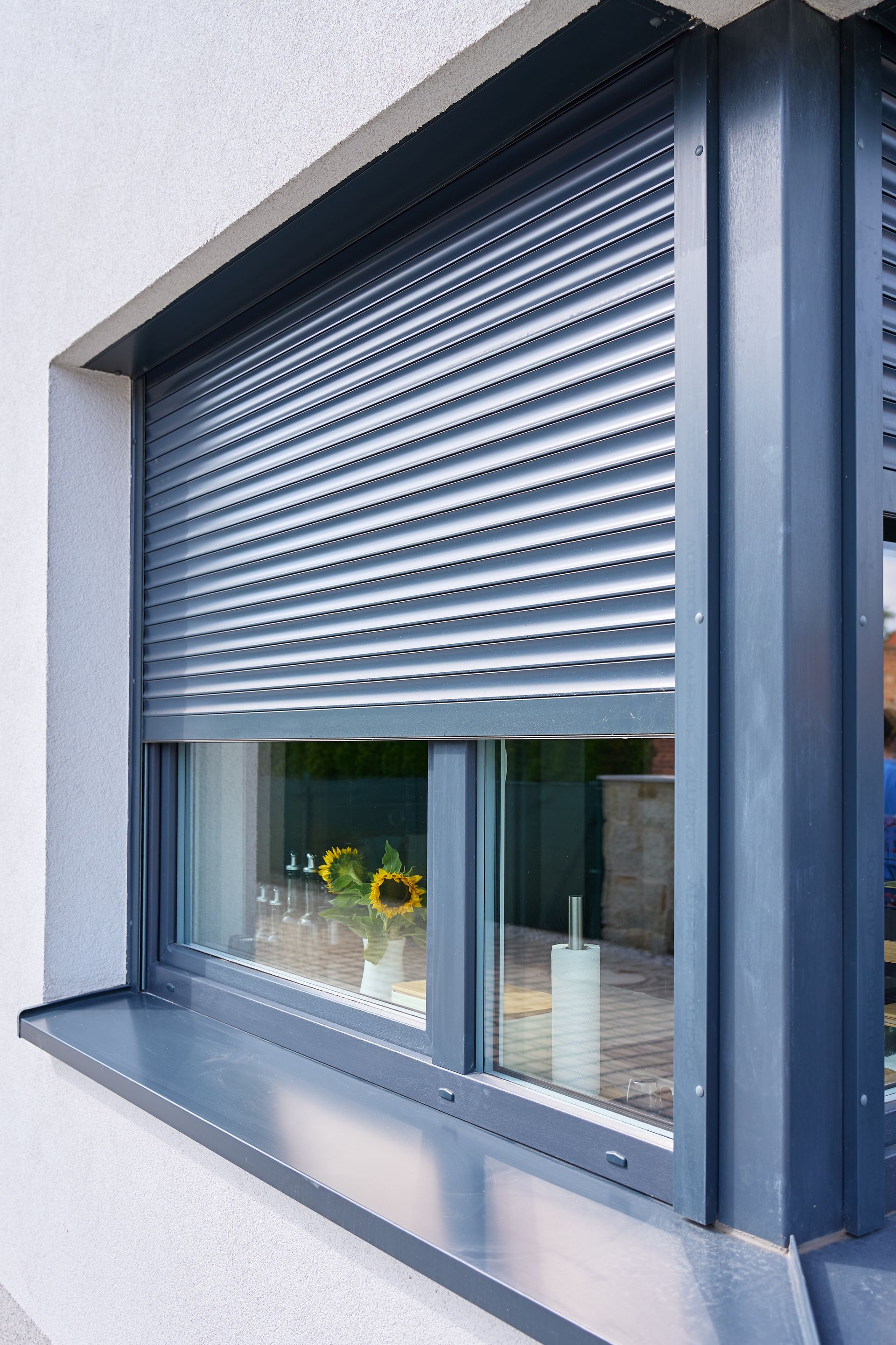 Partially open gray window with a rolled-down shutter and flowers on the sill