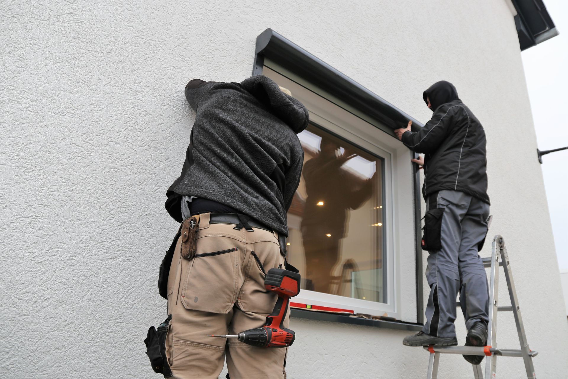 Two workers installing a window on a white exterior wall, one on a ladder and one on a platform