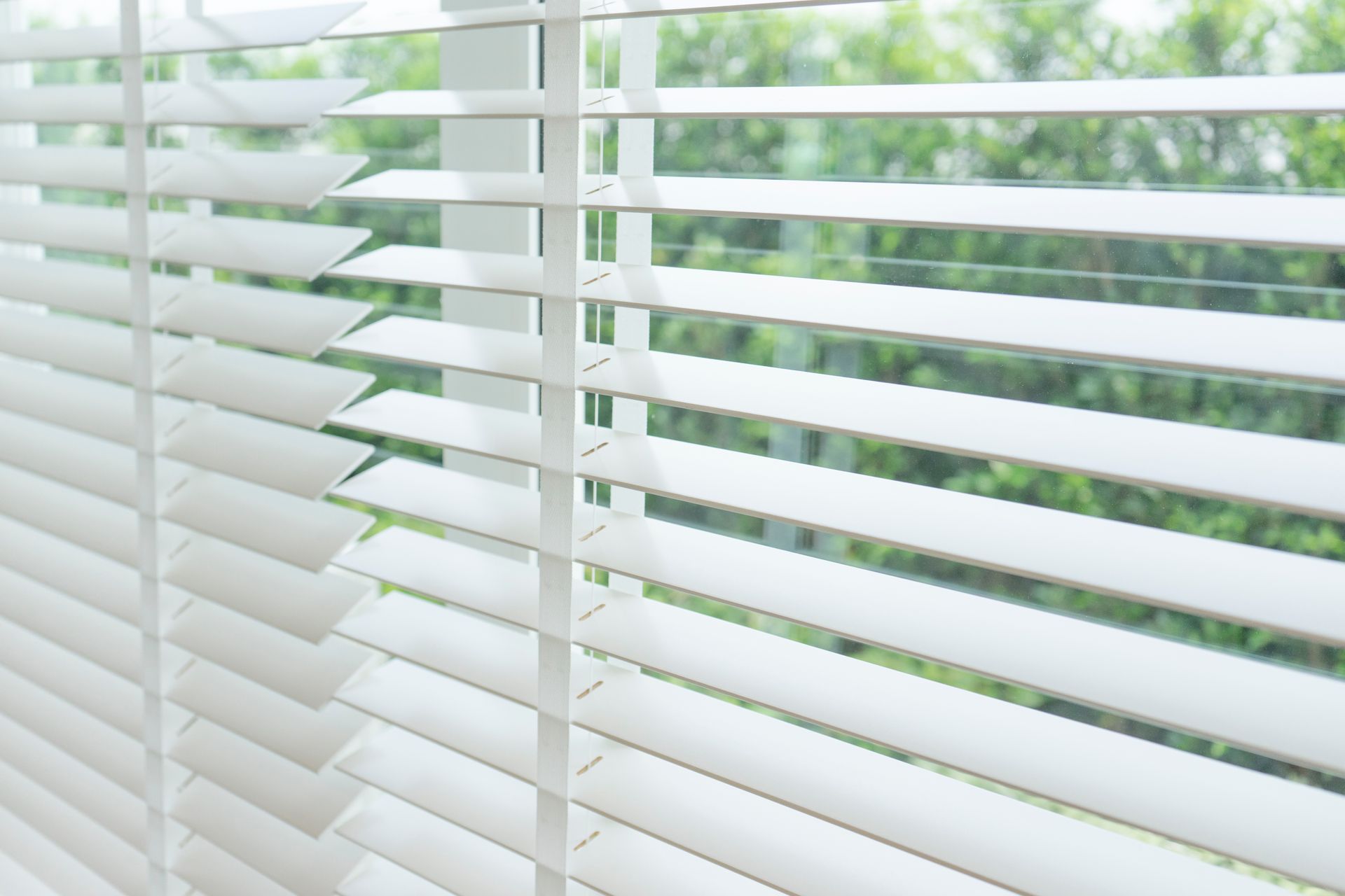 White window blinds with sunlight filtering through, showing a blurred green view outside