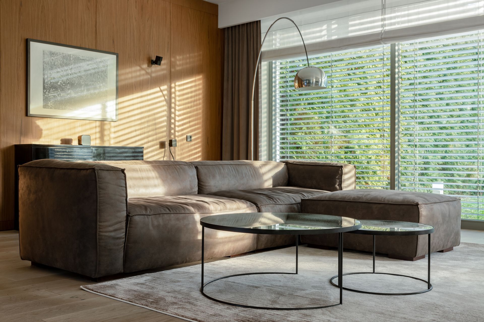 Modern living room with brown sectional sofa, glass coffee tables, wood wall paneling, and bright window light