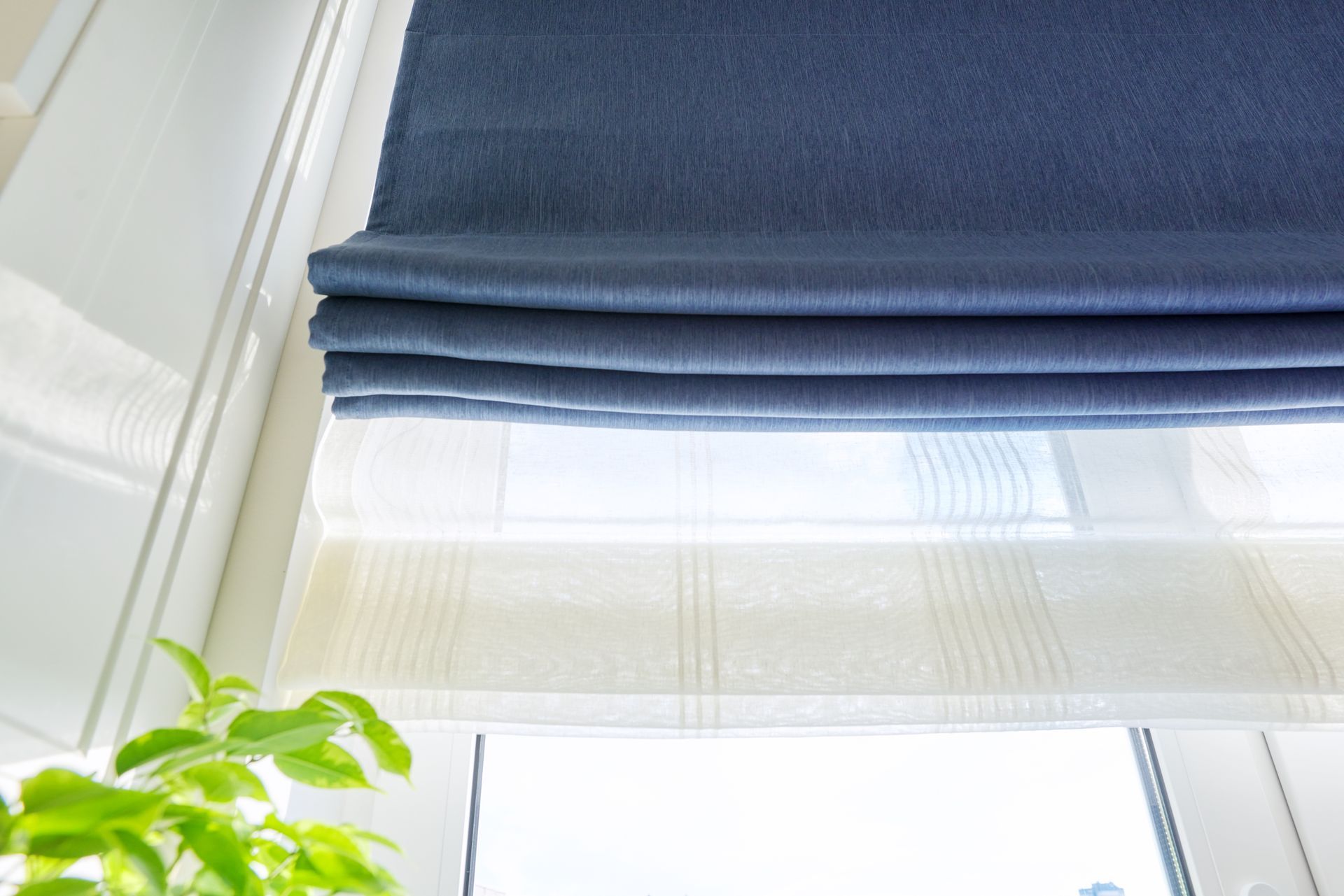 Blue Roman shades above a bright window with a white sheer blind and green plant leaves below