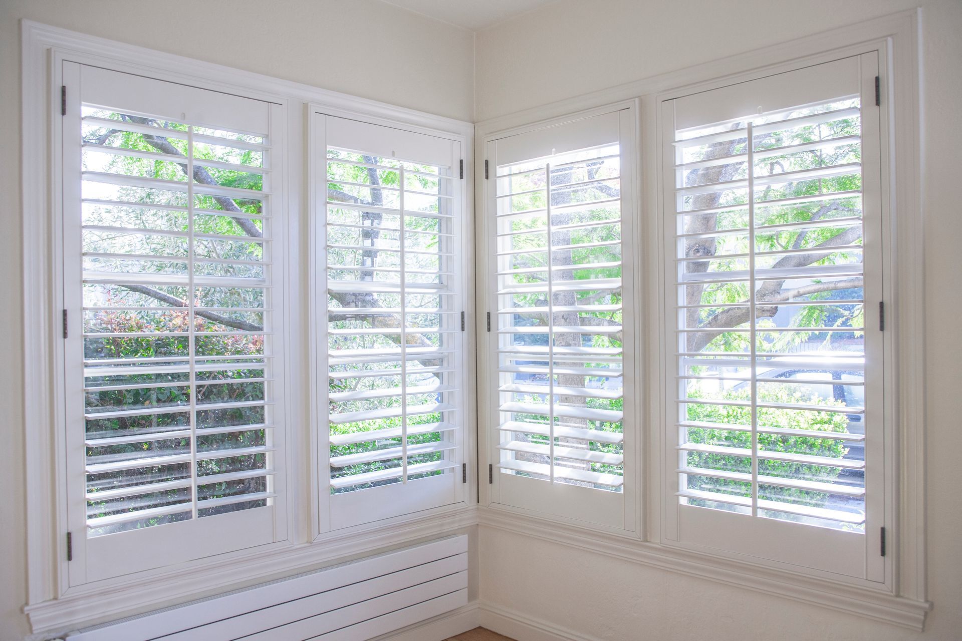 Bright white room corner with large windows and white plantation shutters, showing greenery outside