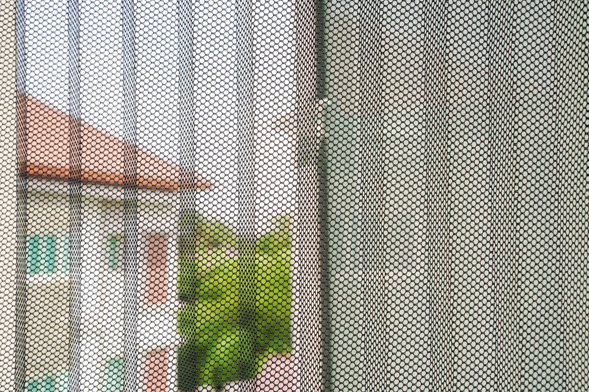View through a screen: white house with red roof and green trees beside a gray wooden wall