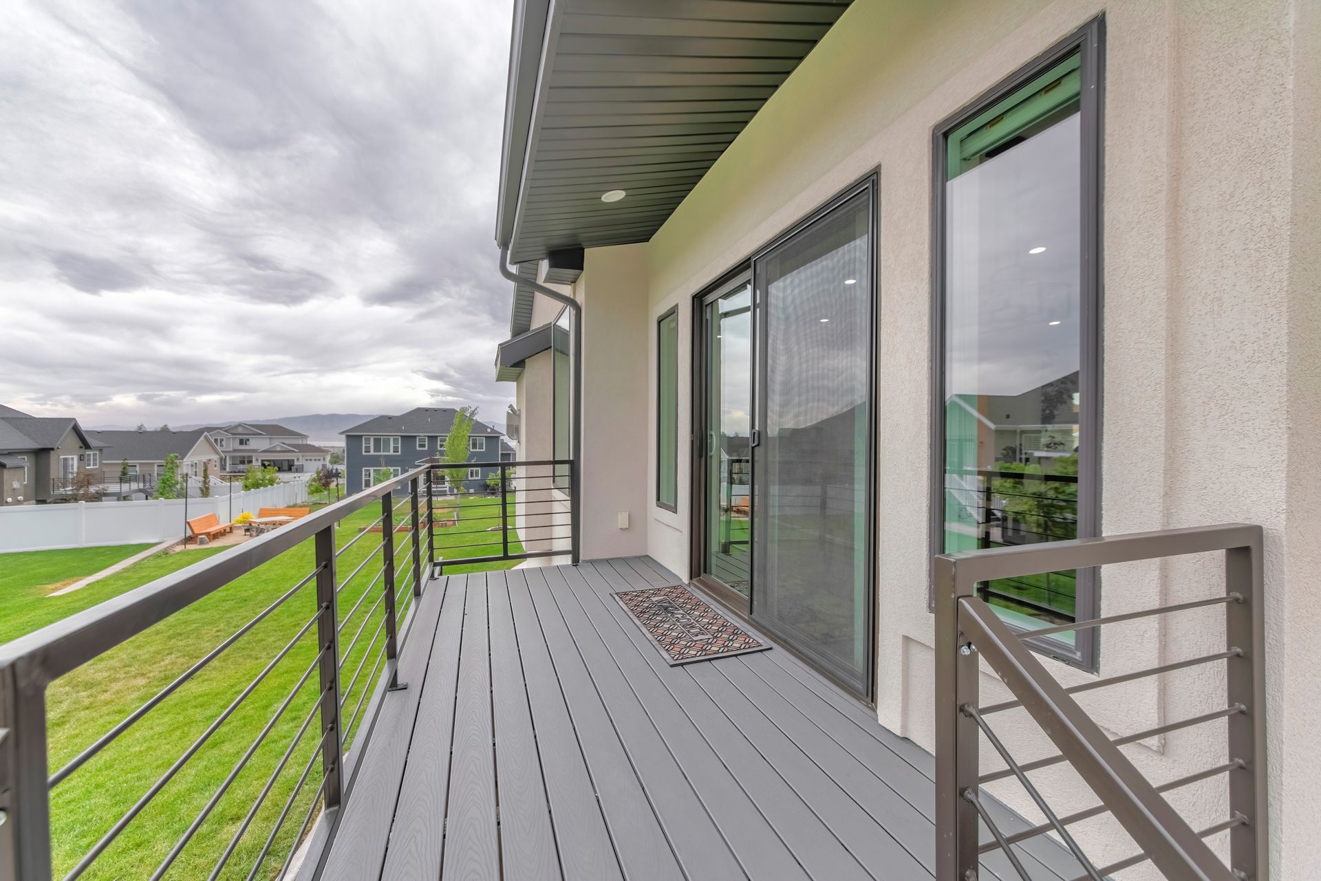 Apartment balcony with gray deck, black railing, and sliding glass doors overlooking a green yard and neighborhood