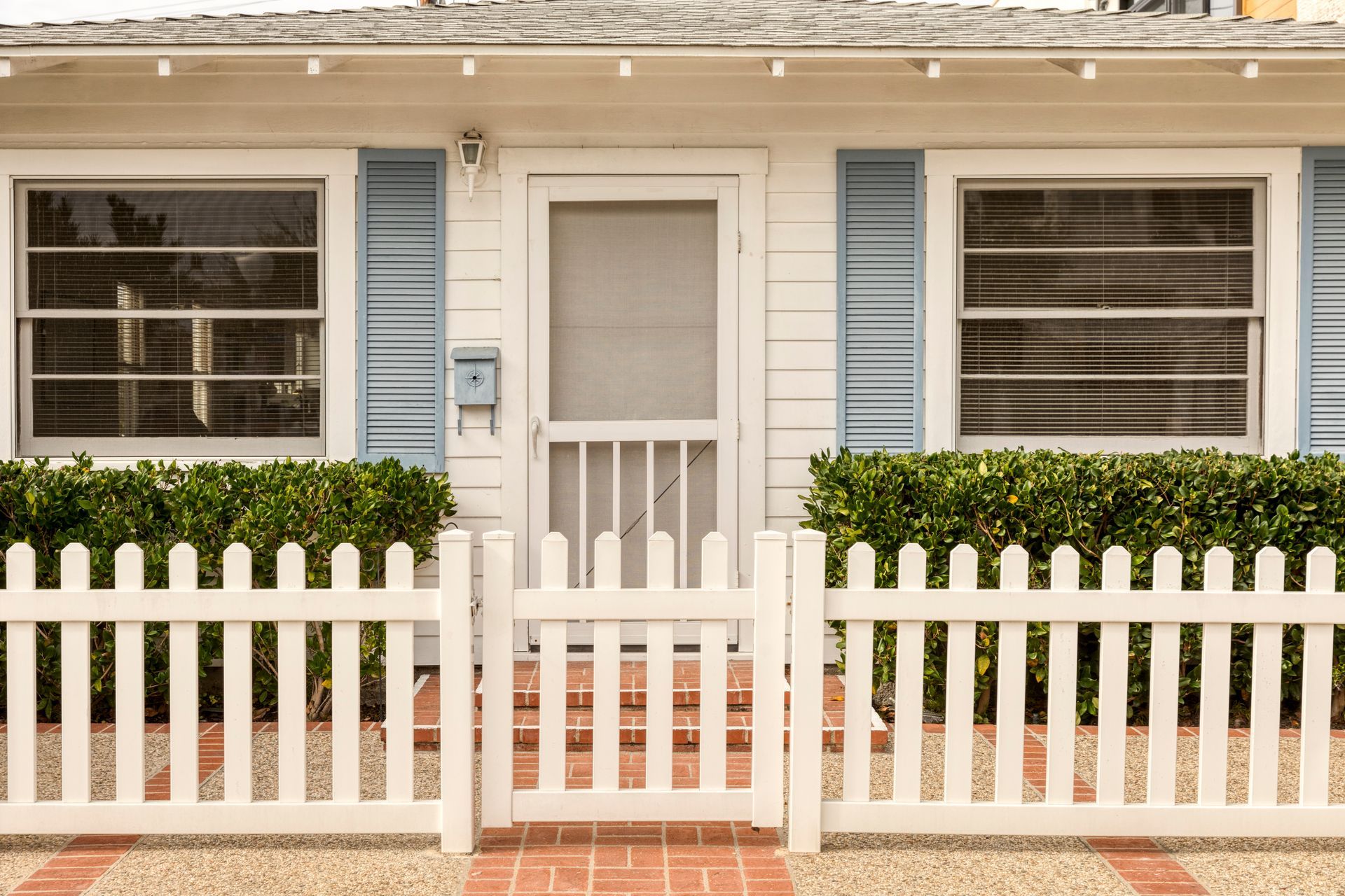 White house front with blue shutters, screened door, and white picket fence in front of hedges