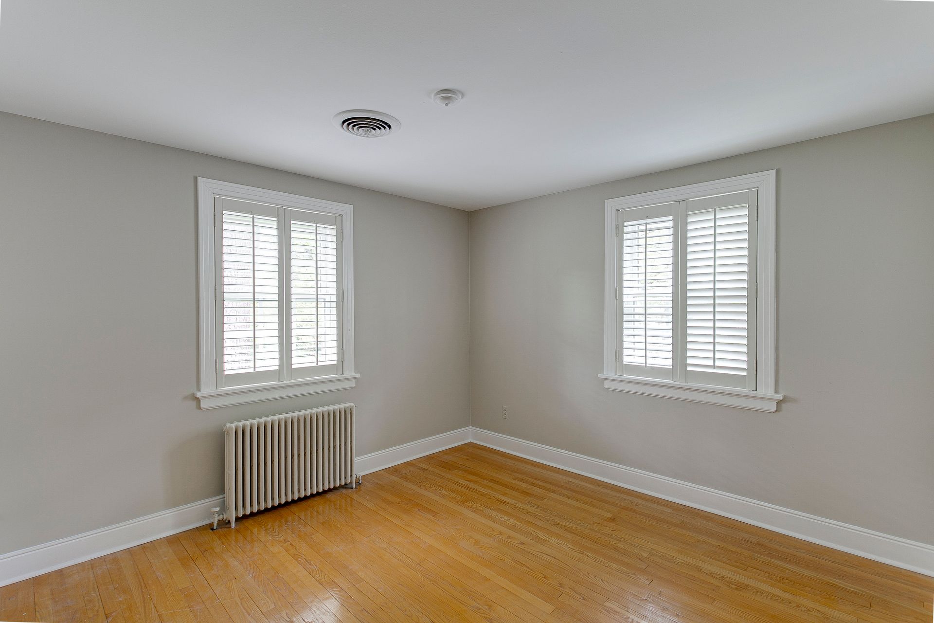Empty beige room with hardwood floor, two windows with white blinds, and a radiator under one window